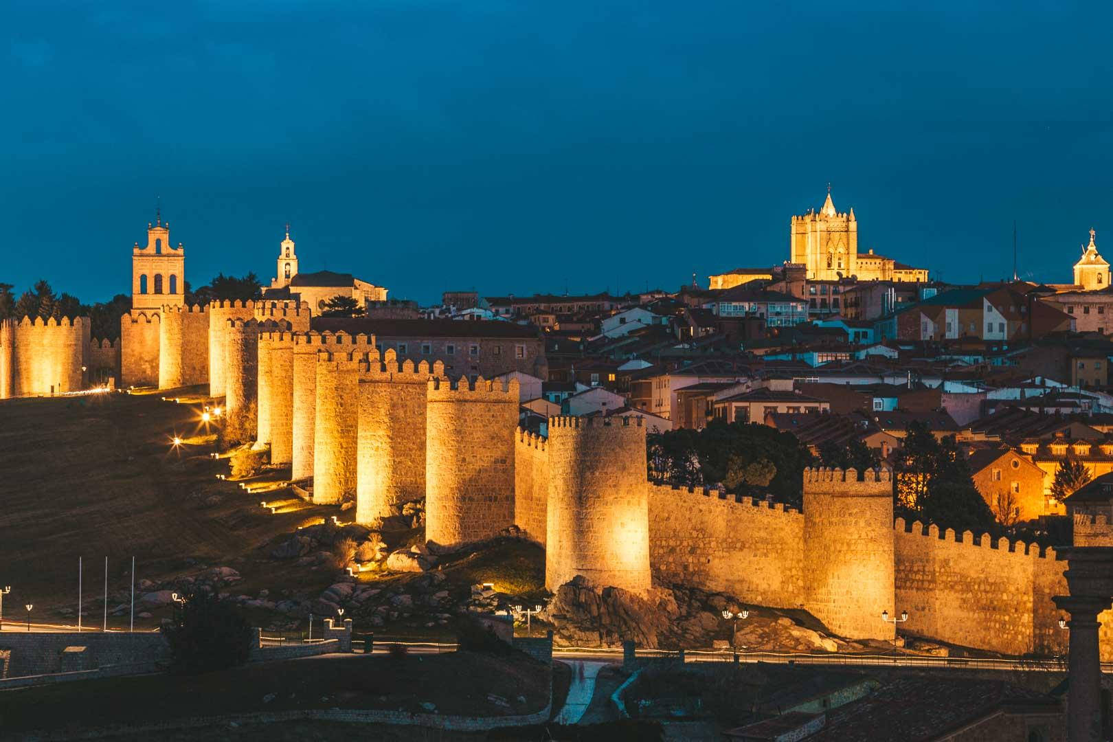 Madrid 桌布 壁紙 — 古老的石牆在暮色燈光下溫暖地發光，環繞著一個歷史悠久的小鎮，教堂尖塔映襯著深藍色的天空。
