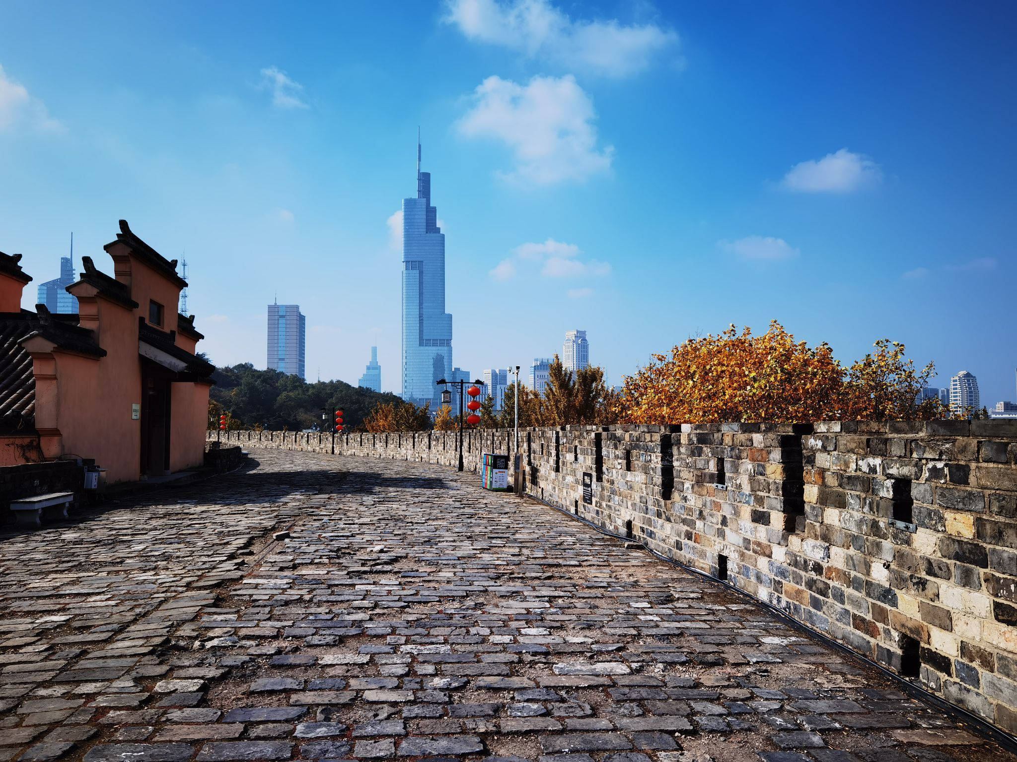 Nanjing 桌布 壁紙 — 古老的石牆小徑與傳統大門通往現代摩天大樓，天空湛藍，雲朵點綴。