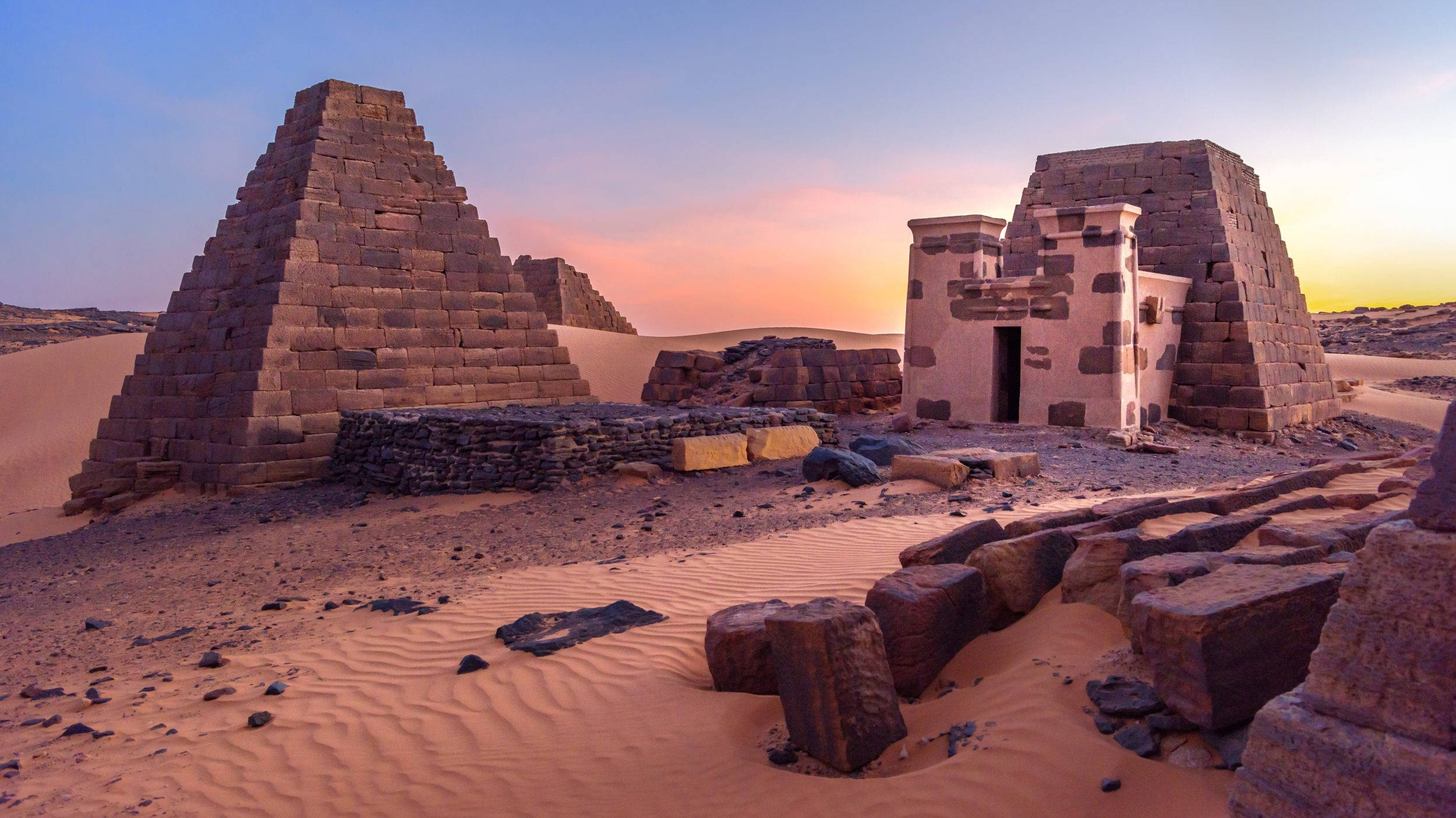 Sudan 桌布 壁紙 — 古老的石造金字塔和寺廟在沙漠日落時分，在溫暖的橙色和粉紅色天空下莊嚴地聳立。