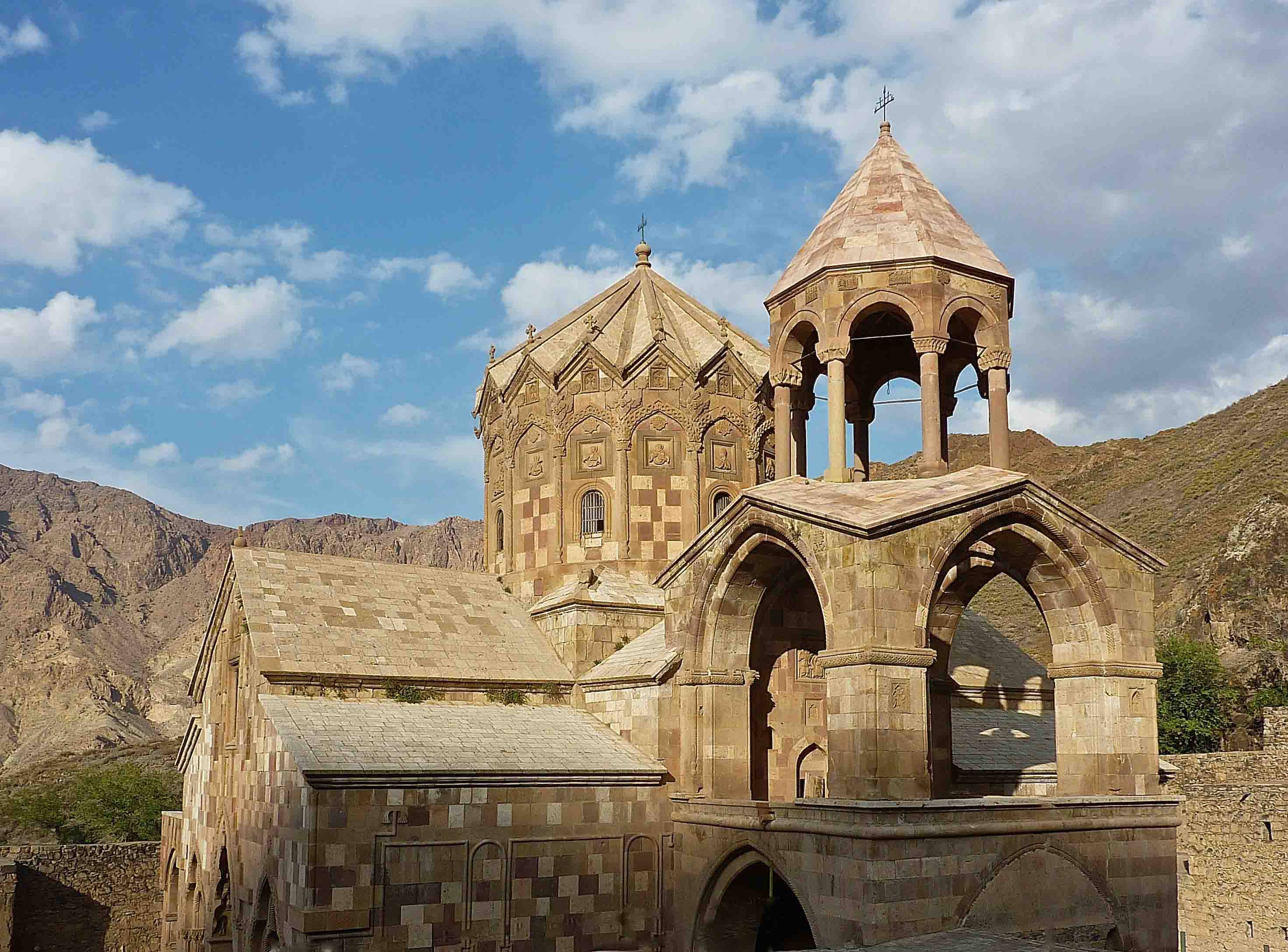 Iran 桌布 壁紙 — 古老的石頭修道院，帶有拱形塔樓和圓頂，在明亮的藍天白雲下，雄偉地矗立在崎嶇的山脈前。