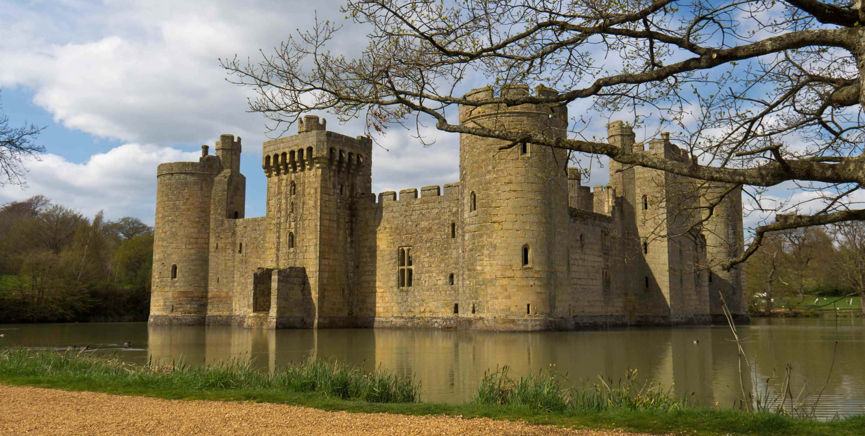 Medieval 桌布 壁紙 — 古老的石頭城堡帶有塔樓，在平靜的護城河中雄偉地矗立，部分多雲的天空被光禿禿的樹枝框住。