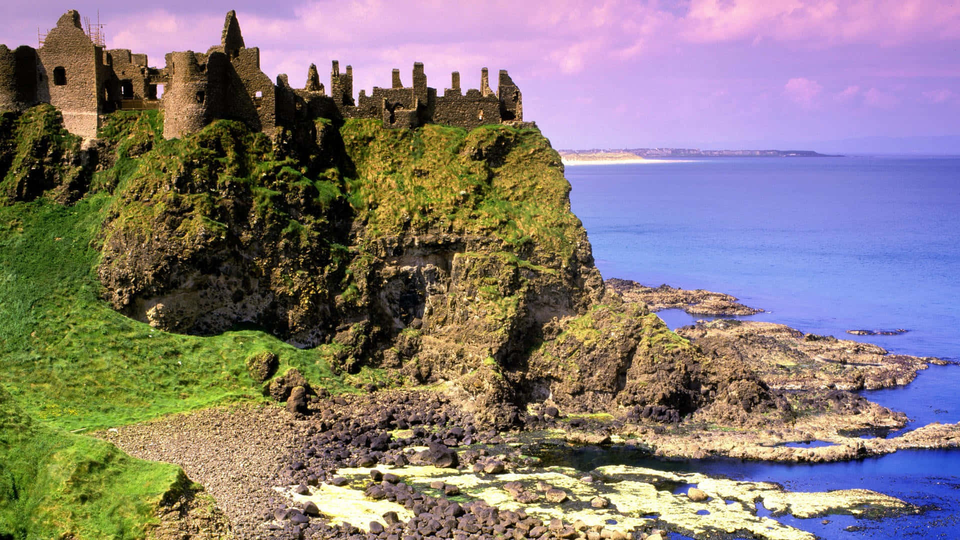 Ireland Desktop 桌布 壁紙 — 古老的石頭城堡廢墟戲劇性地坐落在一個長滿苔蘚的懸崖上，俯瞰著紫色天空下的廣闊藍色大海。