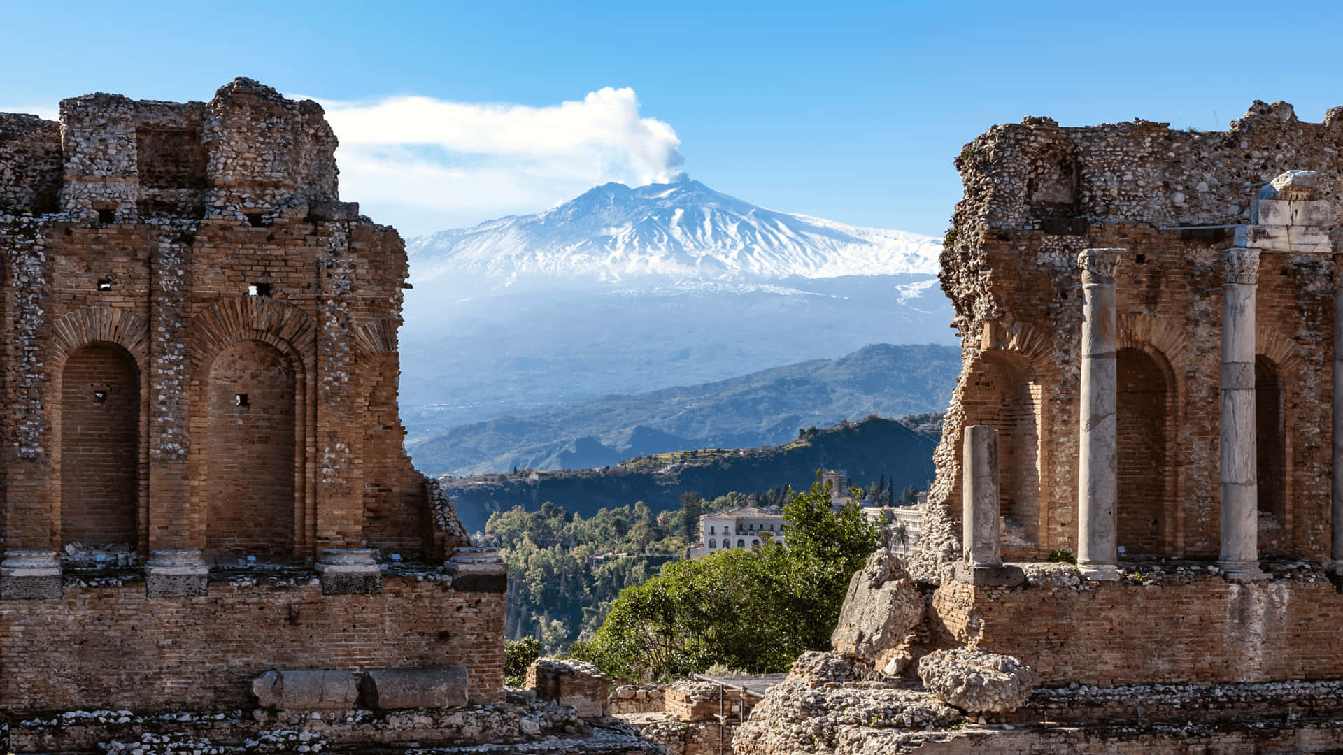 山景 桌布 壁紙 — 古老的石頭廢墟框住一座雄偉的雪山火山，背景是晴朗的藍天和飄浮的白雲。