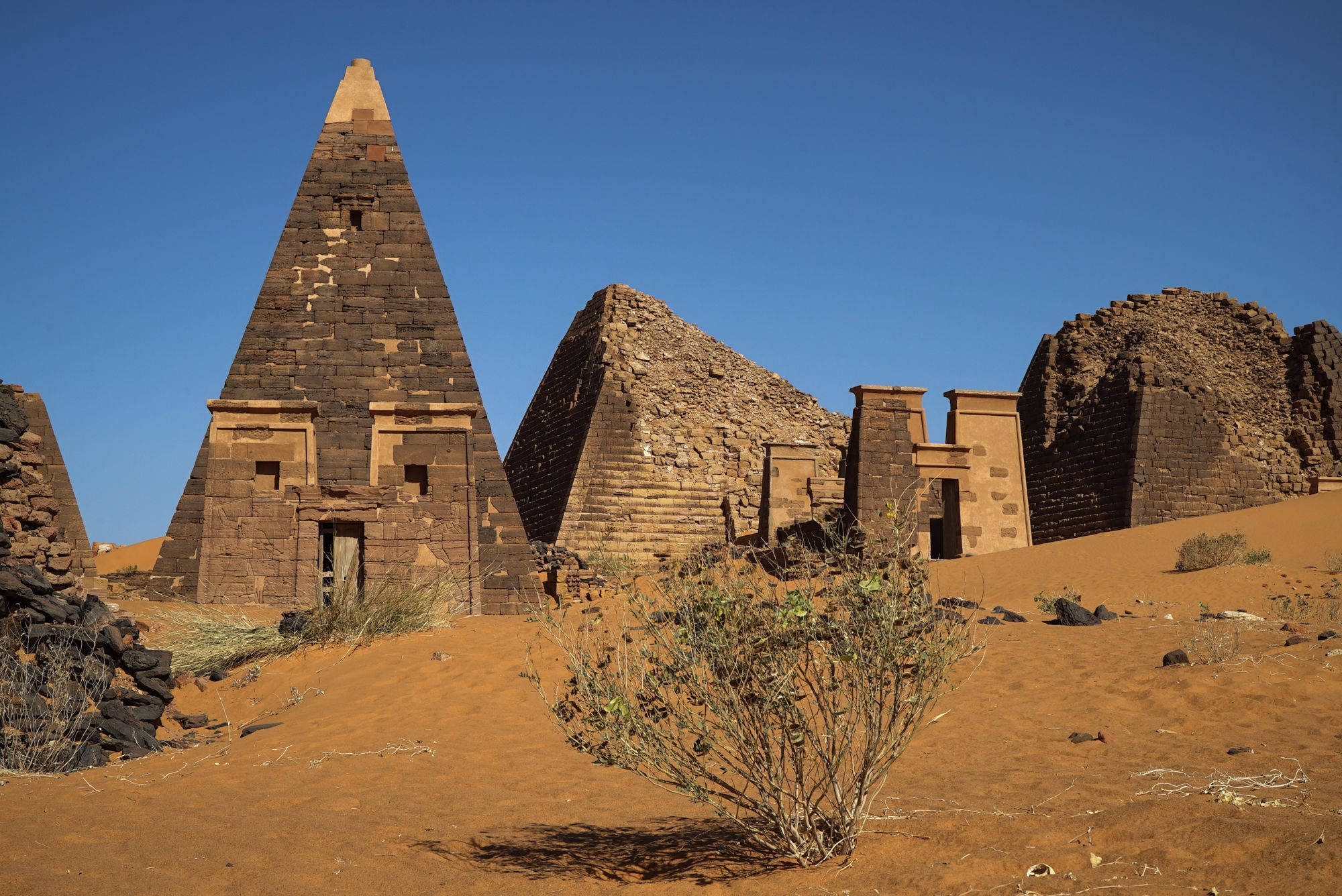 Sudan 桌布 壁紙 — 古老的石頭金字塔和廢墟在廣闊的沙漠中，在晴朗的藍天下莊嚴地聳立，周圍有稀疏的乾燥植物。