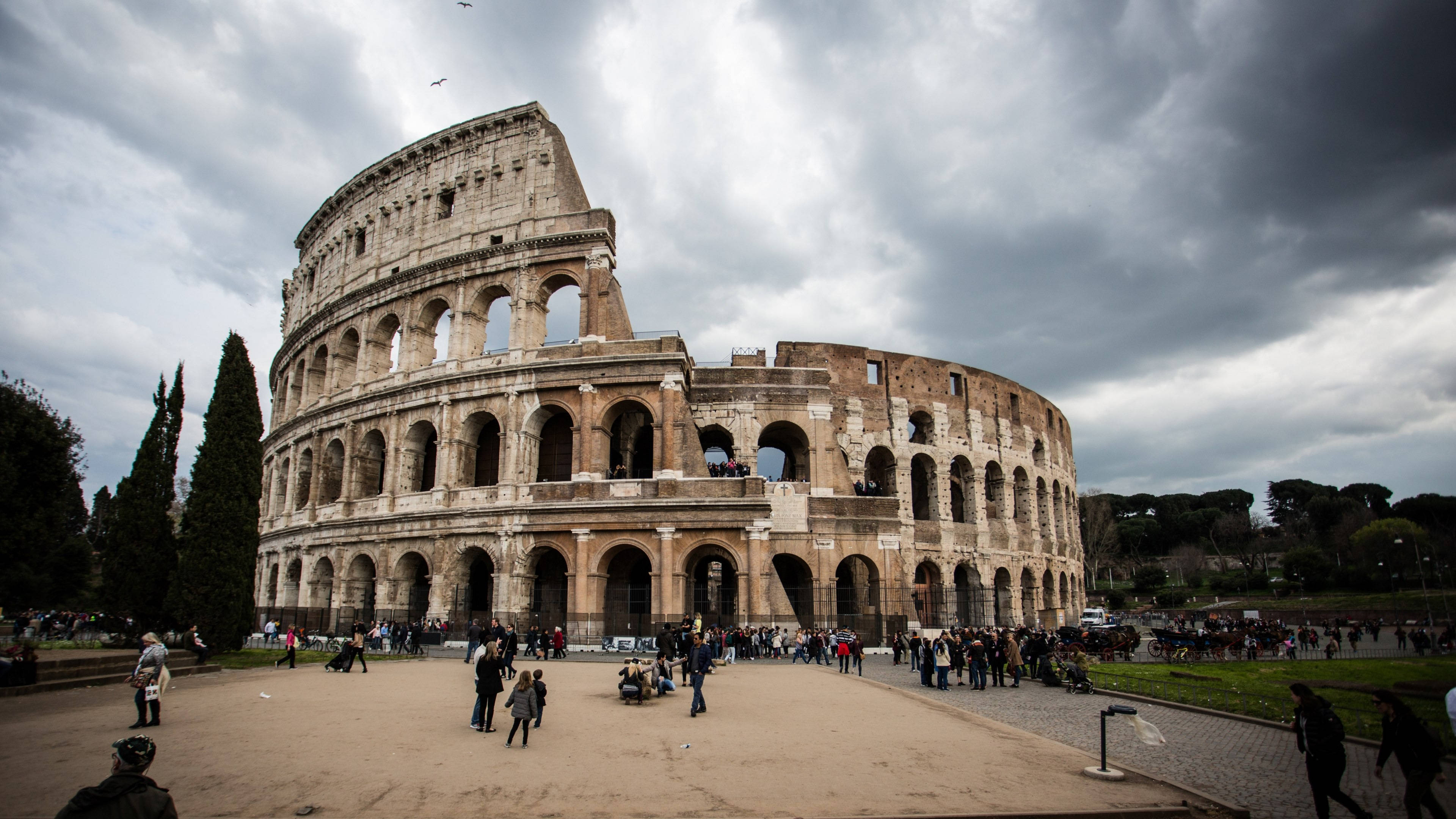 Rome 桌布 壁紙 — 古老的羅馬競技場在戲劇性的多雲天空下矗立，遊客正在探索其巨大的石拱門和風化的外牆。
