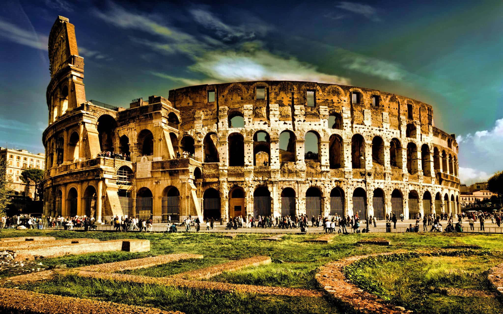 Rome 桌布 壁紙 — 古老的羅馬競技場在戲劇性的天空下雄偉地矗立，遊客正在探索其歷史遺跡和周圍的草地。
