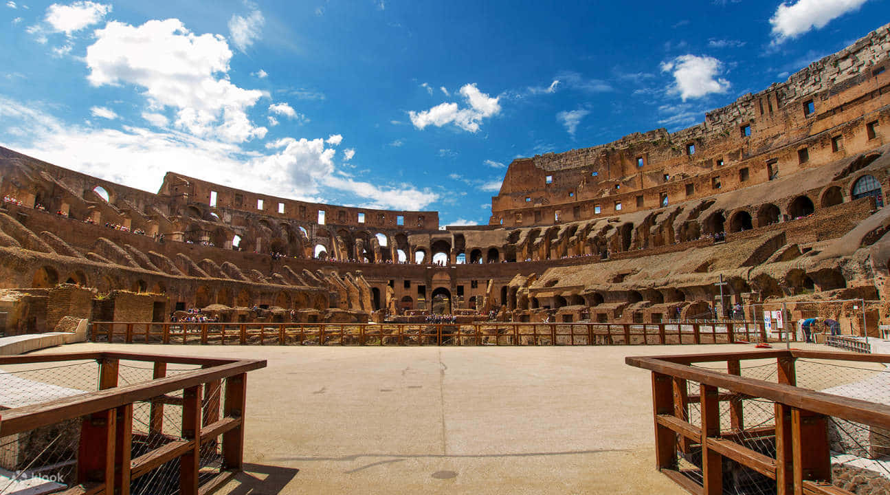 Roman Colosseum 桌布 壁紙 — 古老的羅馬競技場在明亮的藍天下雄偉地矗立，蓬鬆的白雲在其風化的石拱門上投下陰影。