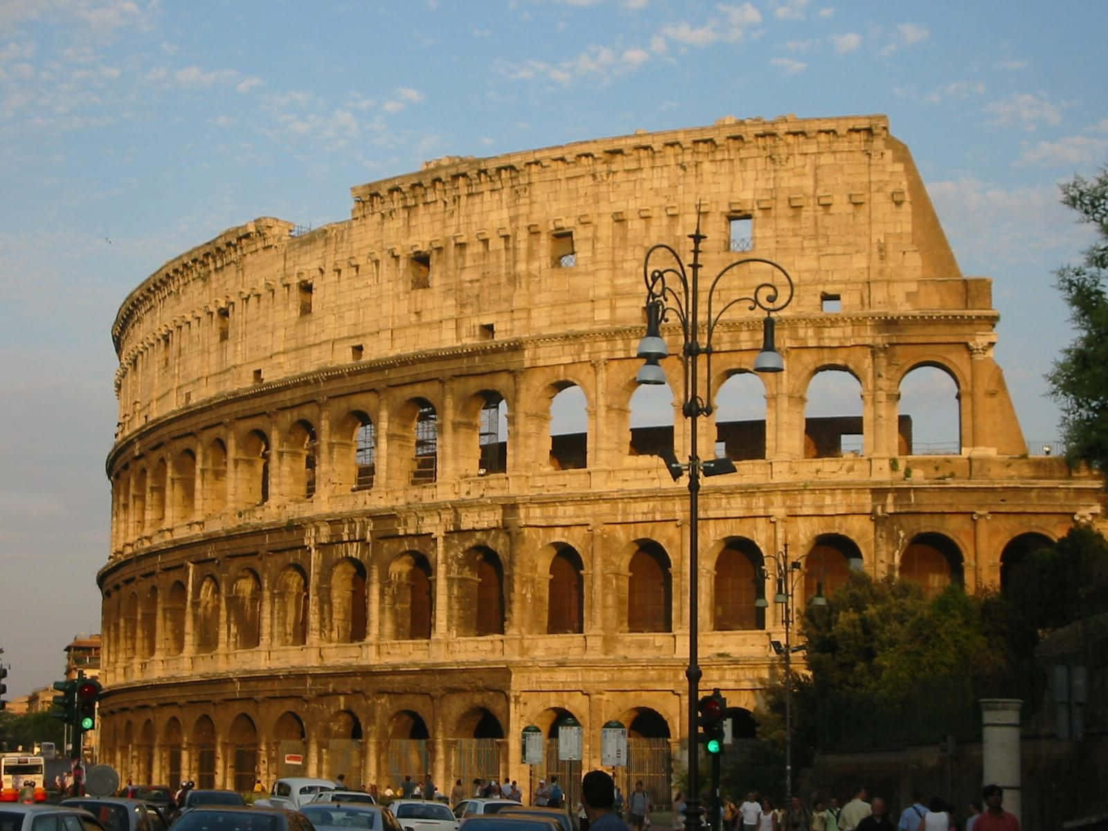 Roman 桌布 壁紙 — 古老的羅馬競技場在柔和藍天與零星白雲下巍然聳立，前景街道上汽車和行人往來穿梭。