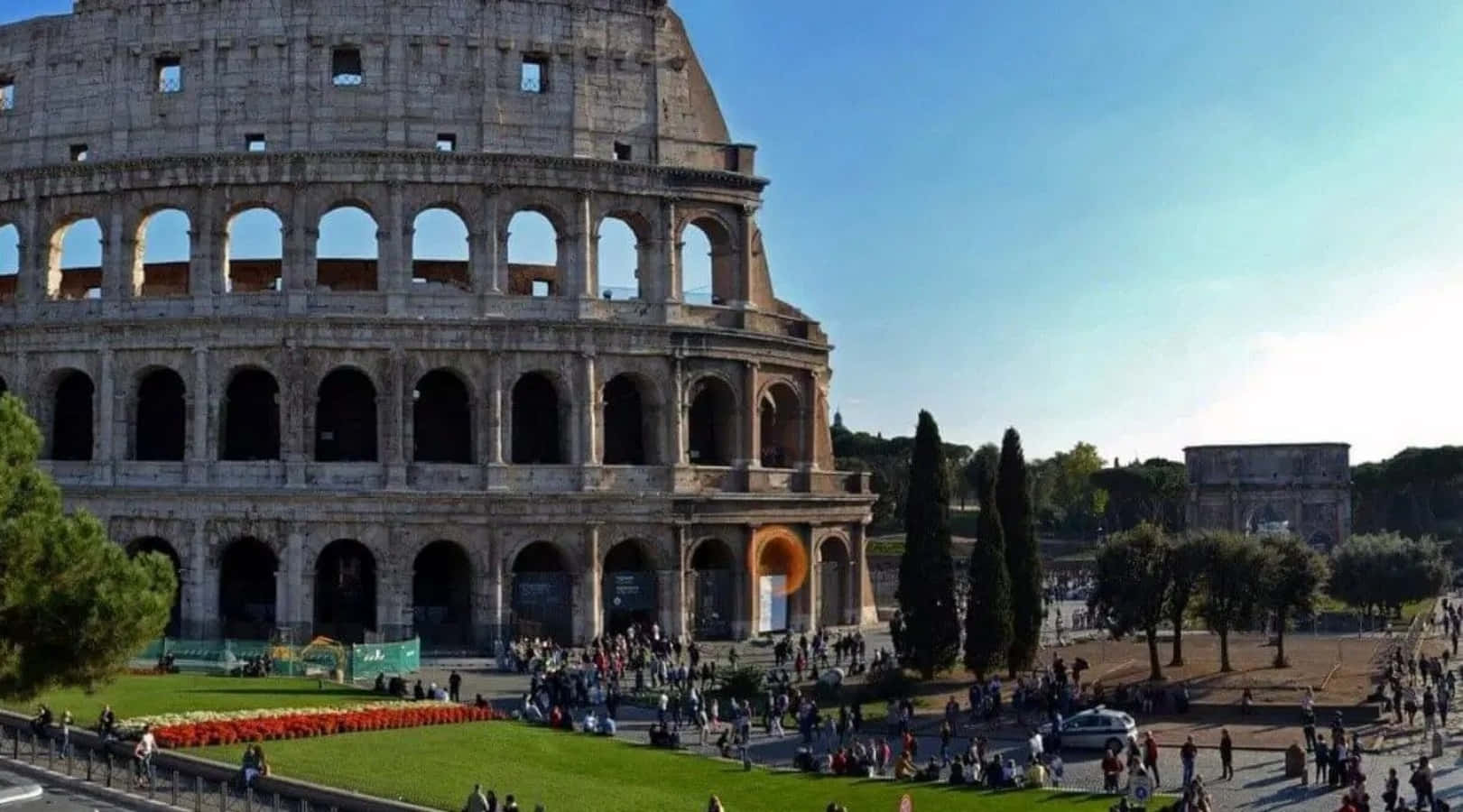 Roman 桌布 壁紙 — 古老的羅馬競技場在清澈的藍天下雄偉矗立，遊客正在探索其歷史遺跡和周圍的綠地。