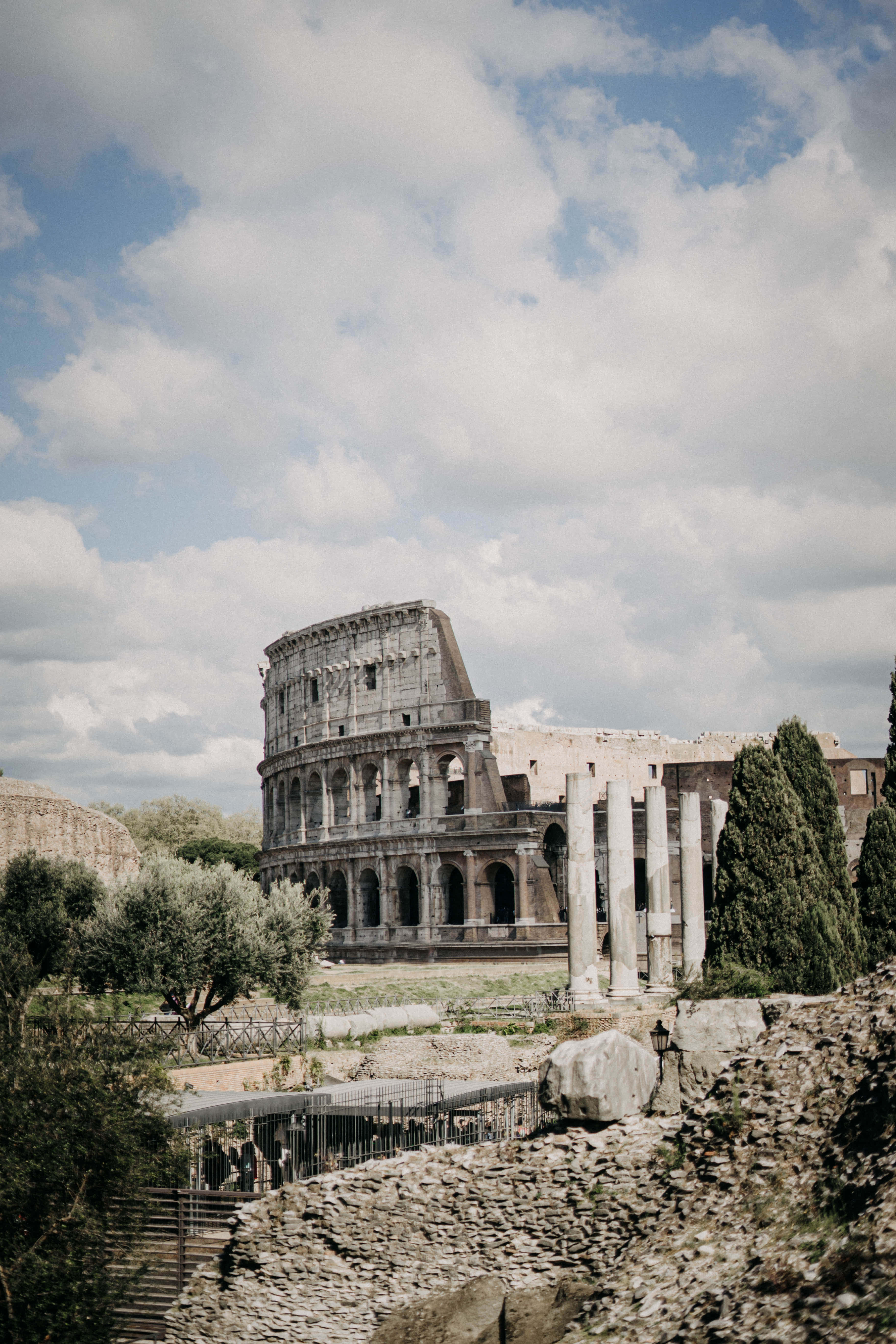 Ancient Roman Desktop 桌布 壁紙 — 古老的羅馬競技場在部分多雲的天空下雄偉地矗立，前景有橄欖樹和廢墟。