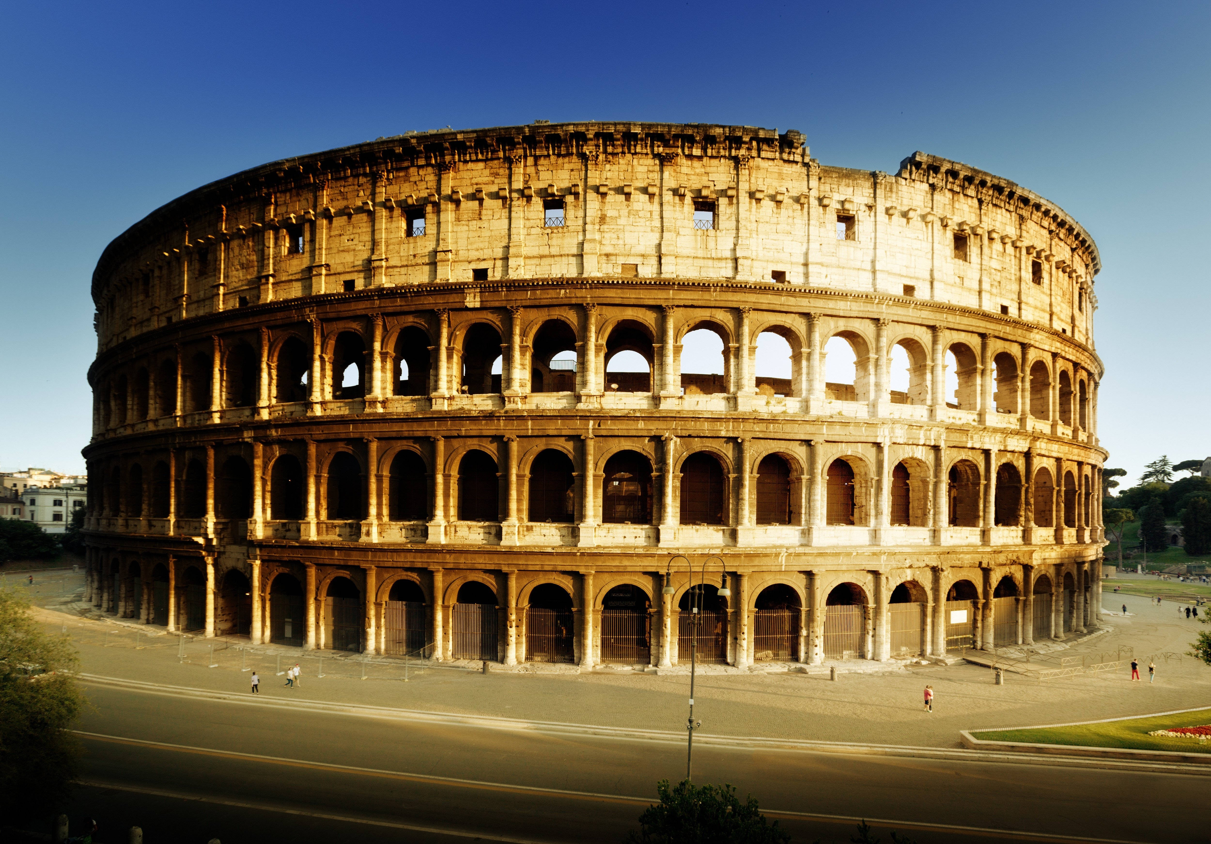 Rome 桌布 壁紙 — 古老的羅馬競技場沐浴在金色的陽光下，天空湛藍，附近有少數遊客走過。