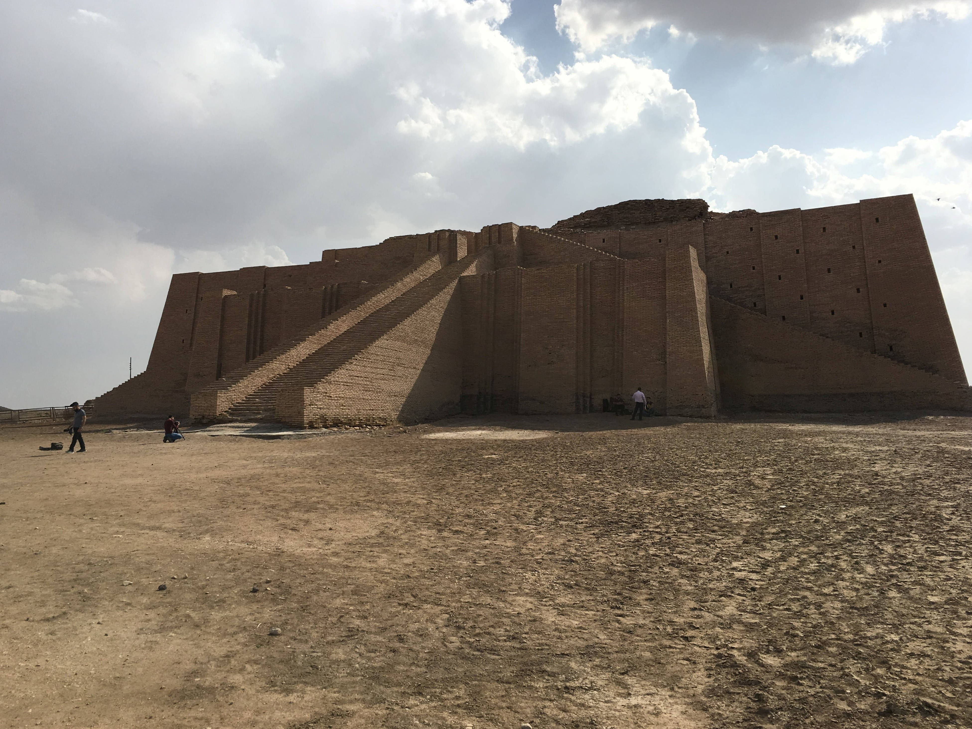 Iraq 桌布 壁紙 — 古老的金字形神廟遺跡從乾旱的沙漠沙地中崛起，天空部分多雲，遊客正在探索其底部。