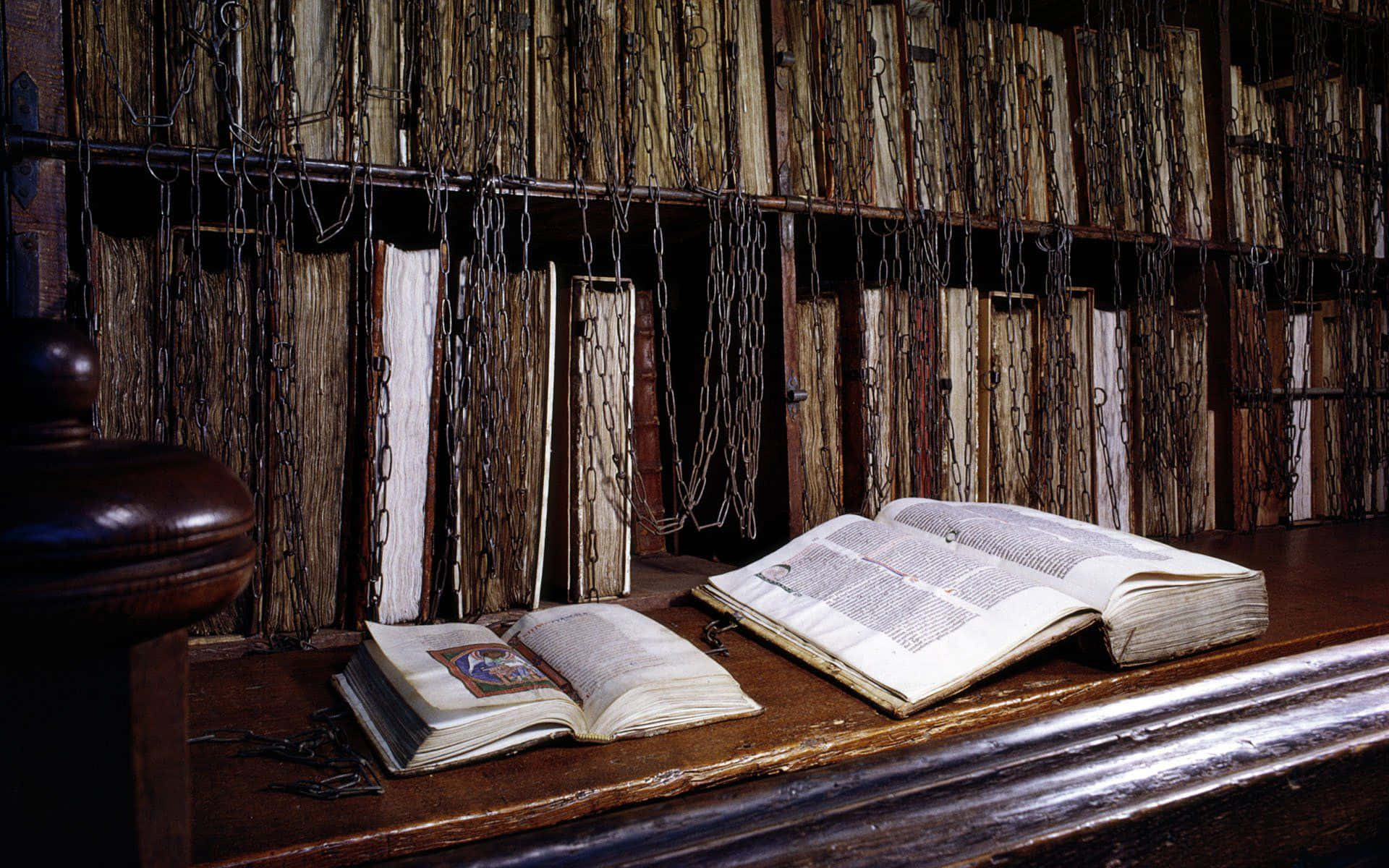 England 桌布 壁紙 — 古老的鎖鏈書本放在木桌上，位於燈光昏暗、書架高聳的圖書館中。