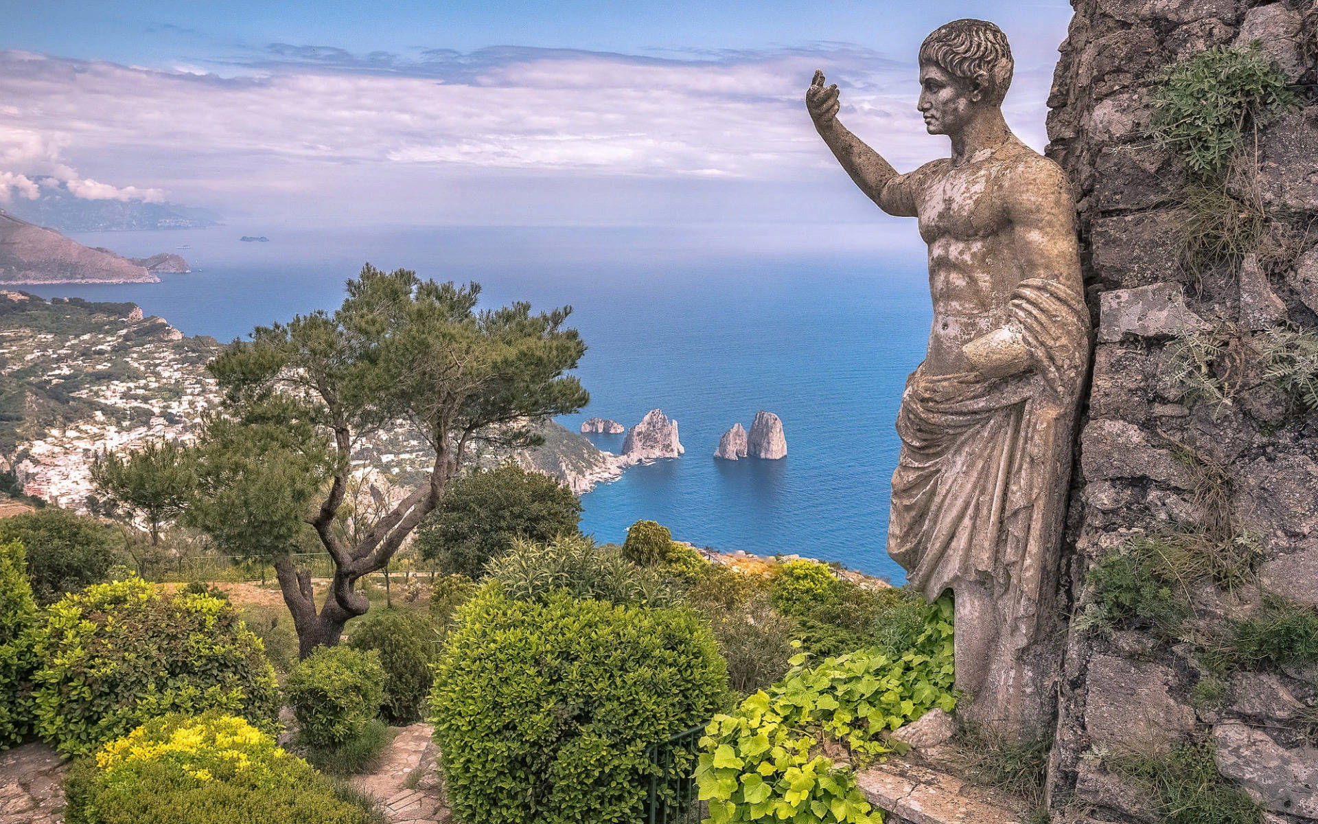 Capri 桌布 壁紙 — 古老石像俯瞰著充滿活力的藍色大海，遠處有懸崖，沿海小鎮坐落在綠色山丘之間。