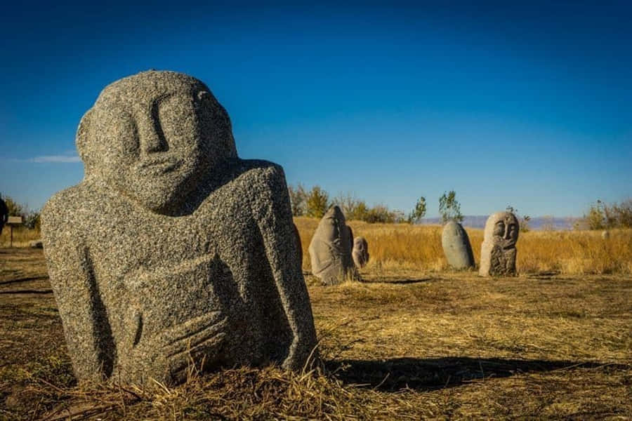 Kyrgyzstan 桌布 壁紙 — 古老石像帶著寧靜的面容，矗立在乾燥的草地上，背景是清澈的藍天。