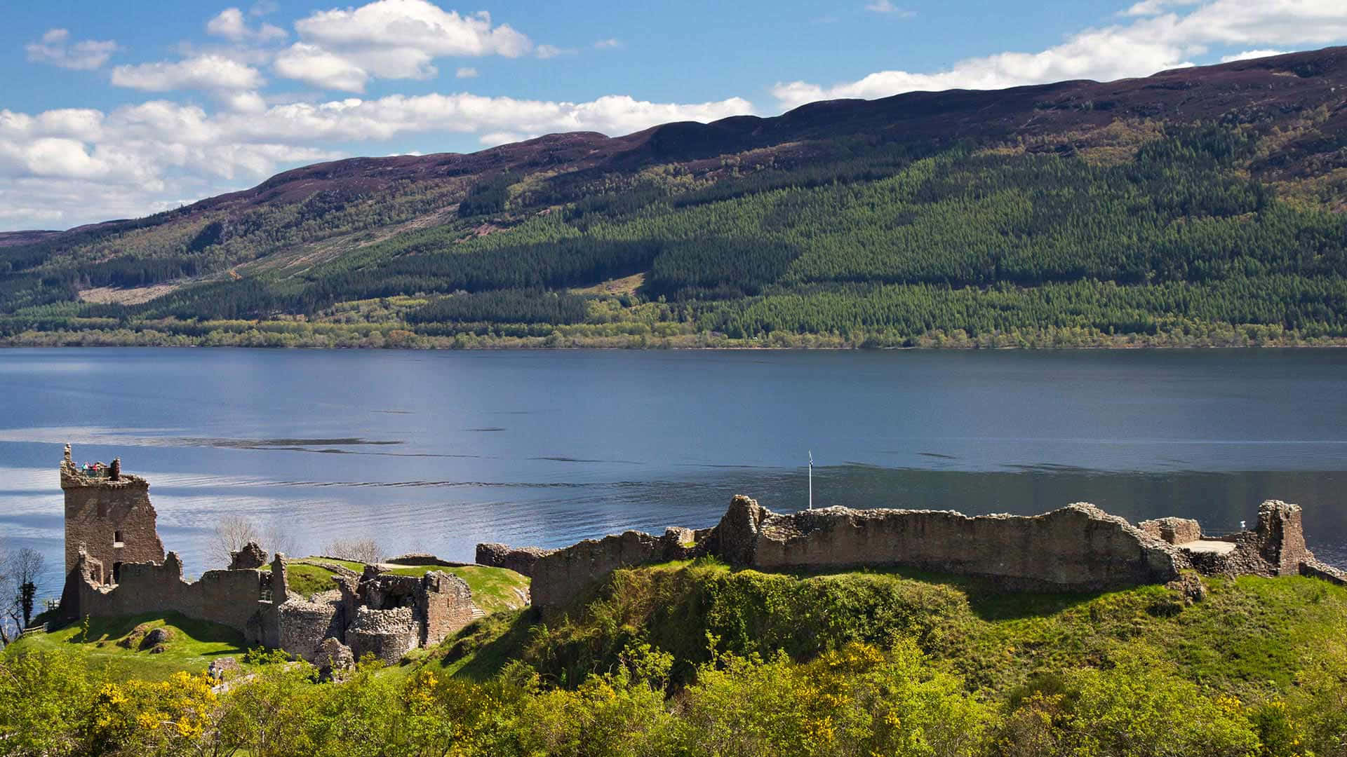 Scotland 桌布 壁紙 — 古老石城堡遺址坐落在長滿草的山丘上，俯瞰著平靜的藍色湖泊，周圍是森林覆蓋的山脈，天空部分多雲。