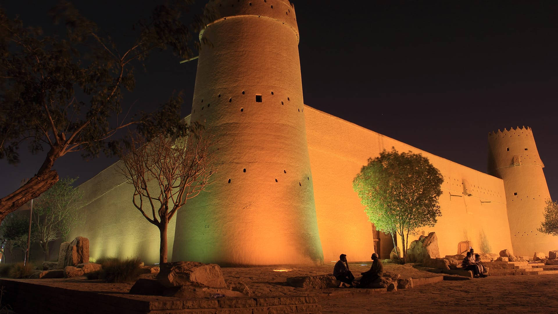 Saudi 桌布 壁紙 — 古老石堡塔在夜燈下溫暖發光，剪影人物在稀疏樹木旁休息。