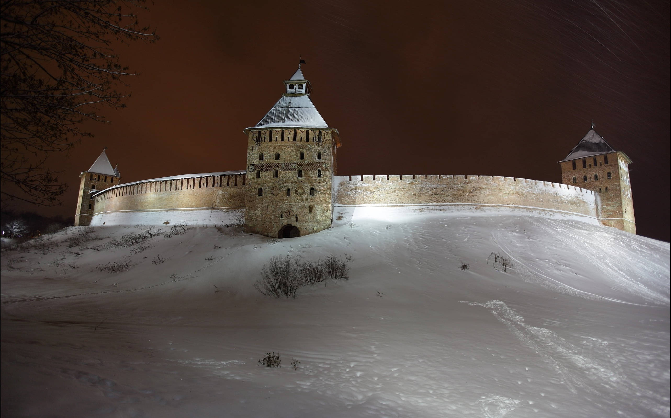 Frozen Castle 桌布 壁紙 — 古老石堡塔從白雪覆蓋的山丘上聳立，在深橙色夜空下飄雪。