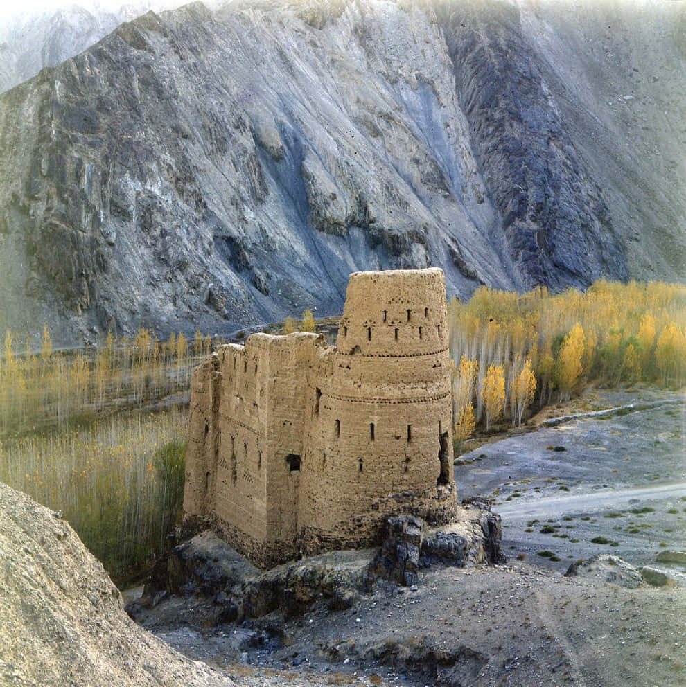 Afghanistan 桌布 壁紙 — 古老石堡孤獨地矗立在河邊，金黃色的秋樹襯托著高聳的灰色山脈。