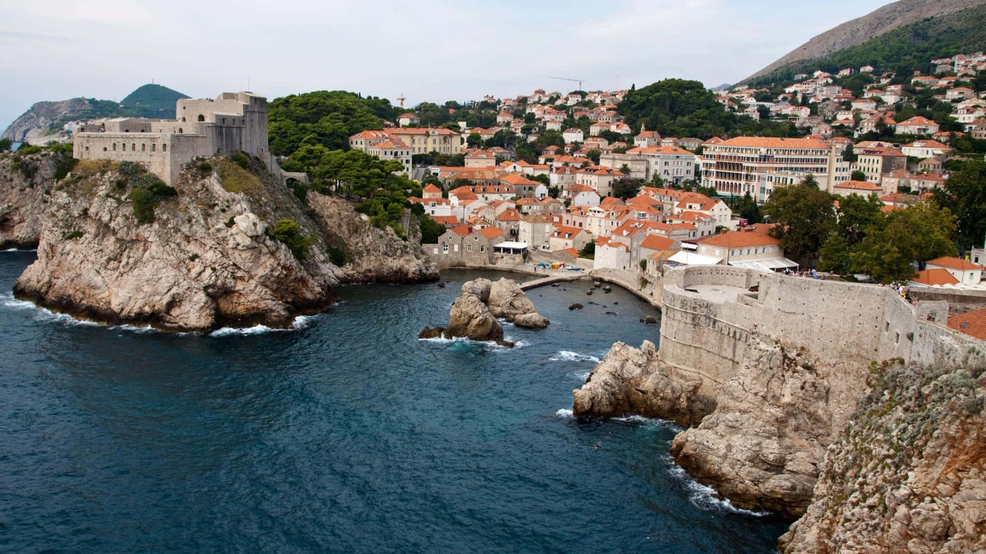 Rocks 桌布 壁紙 — 古老石堡聳立在岩石懸崖上，俯瞰著一個充滿活力的沿海小鎮，鎮上有赤陶屋頂，依偎在深藍色的海邊。