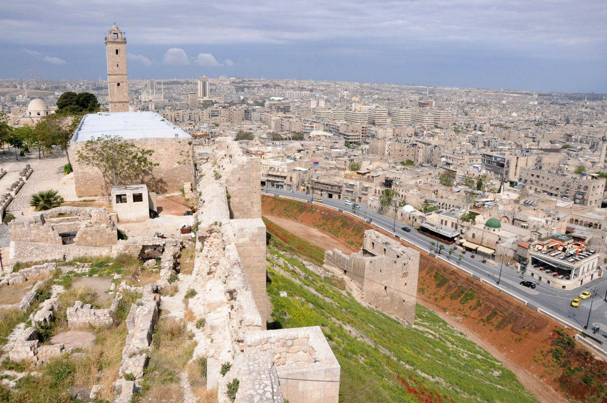 Syria 桌布 壁紙 — 古老石堡遺跡俯瞰著多雲天空下廣闊的現代城市景觀。