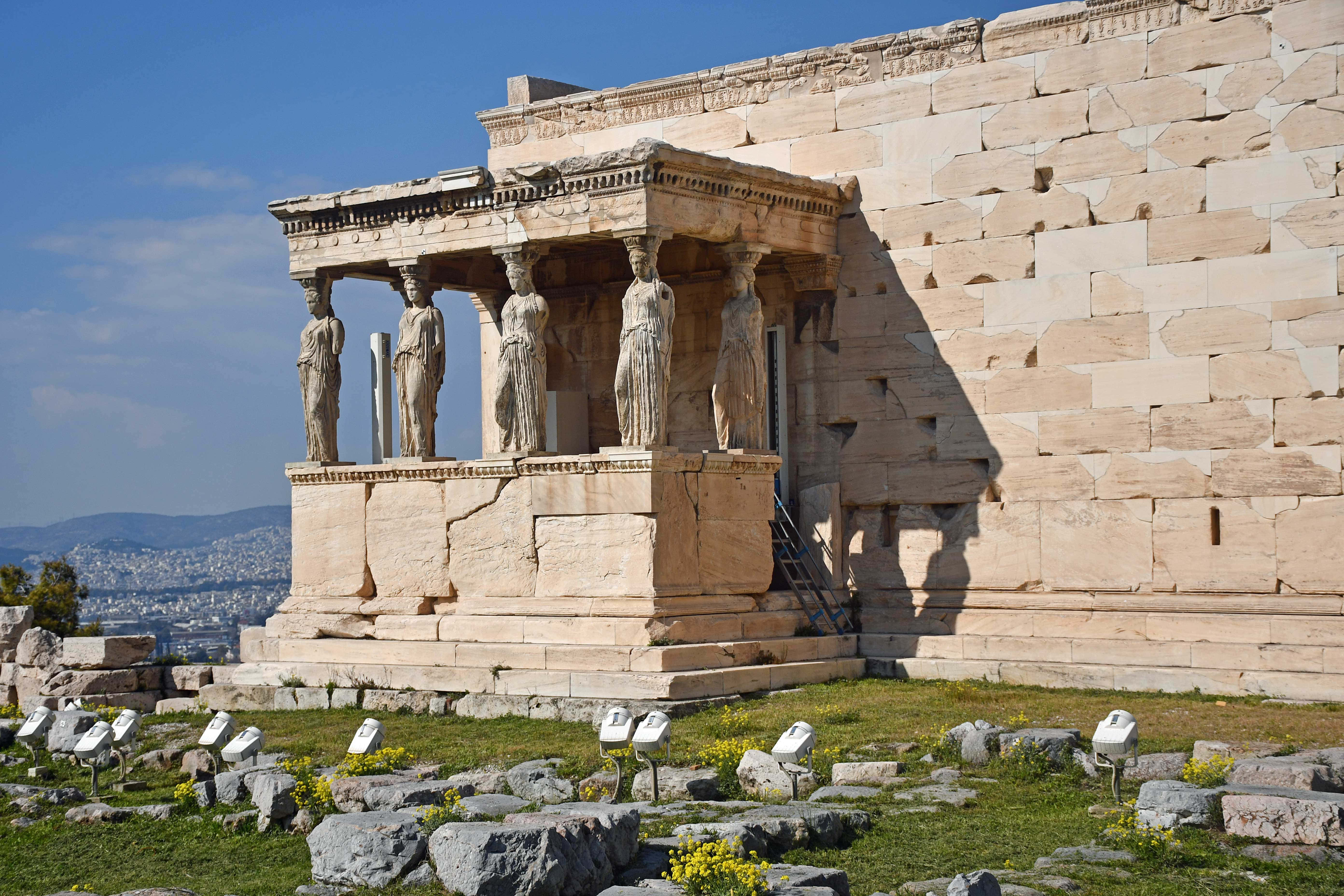 greek 桌布 壁紙 — 古老石廟以女像柱支撐，矗立在明亮的藍天下，俯瞰城市景觀和遠山。