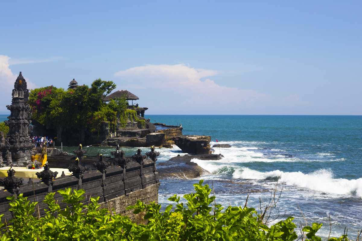 Bali Island 桌布 壁紙 — 古老石廟矗立在海崖上，海浪拍打，綠意盎然，天空湛藍。