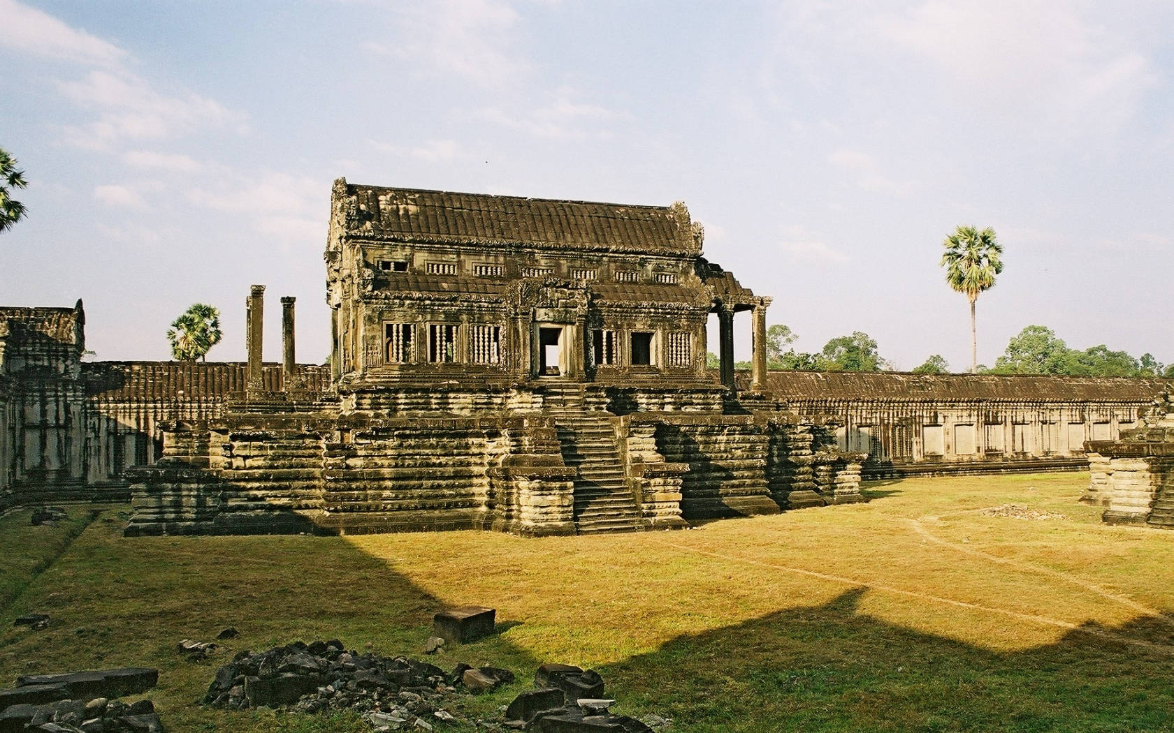 Cambodia 桌布 壁紙 — 古老石廟遺跡，有分層平台和斷裂的柱子，矗立在晴朗的天空下，有零星雲朵。