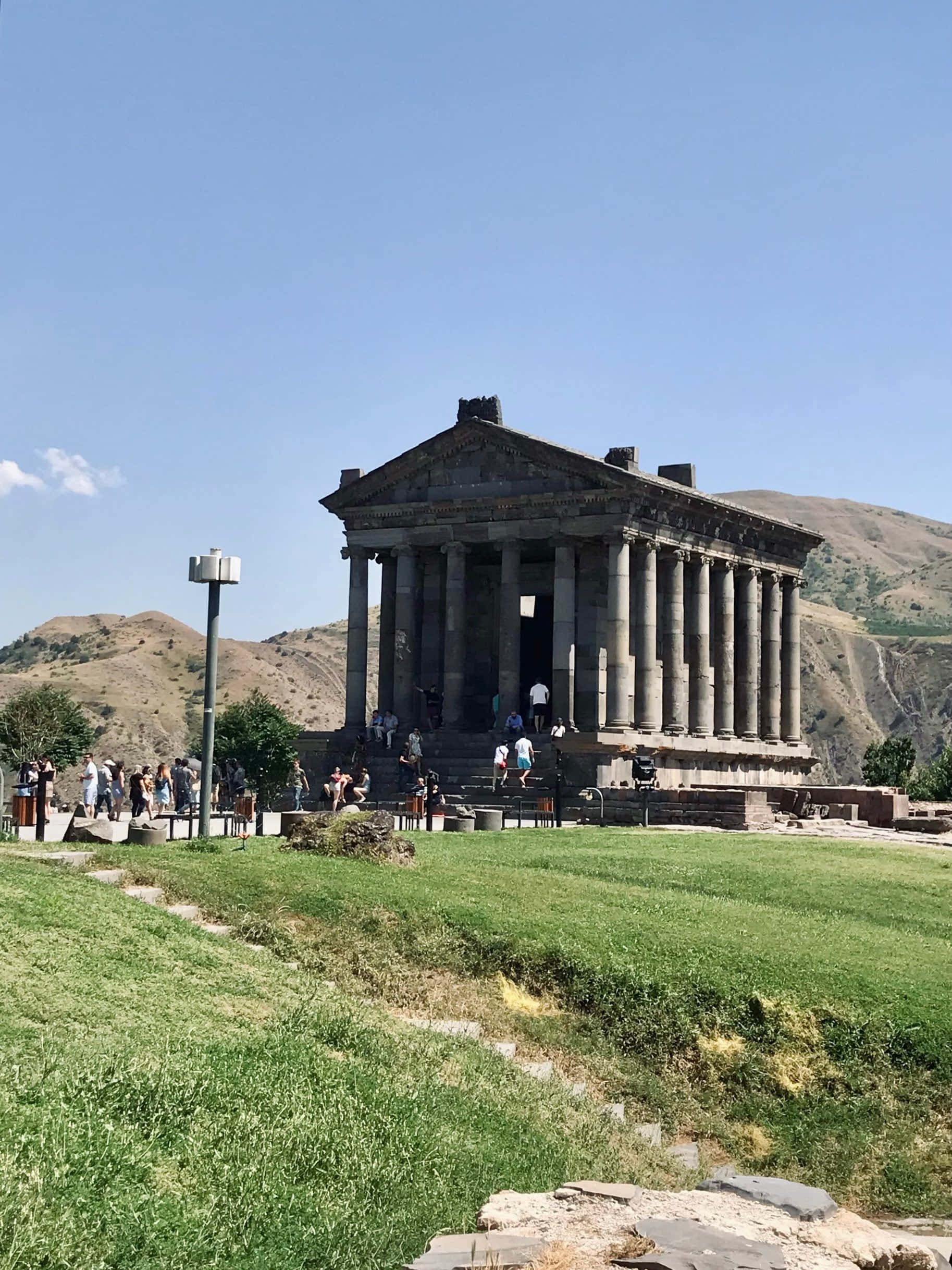 建築 桌布 壁紙 — 古老石廟高柱聳立於青草山丘上，藍天白雲下遊客探索其階梯。