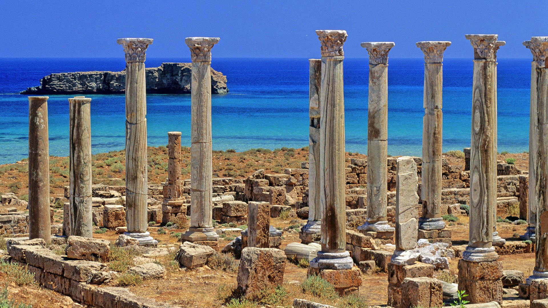 greek 桌布 壁紙 — 古老石柱在廢墟中高聳矗立，俯瞰著清澈藍天下碧綠色的海洋和岩石海岸。