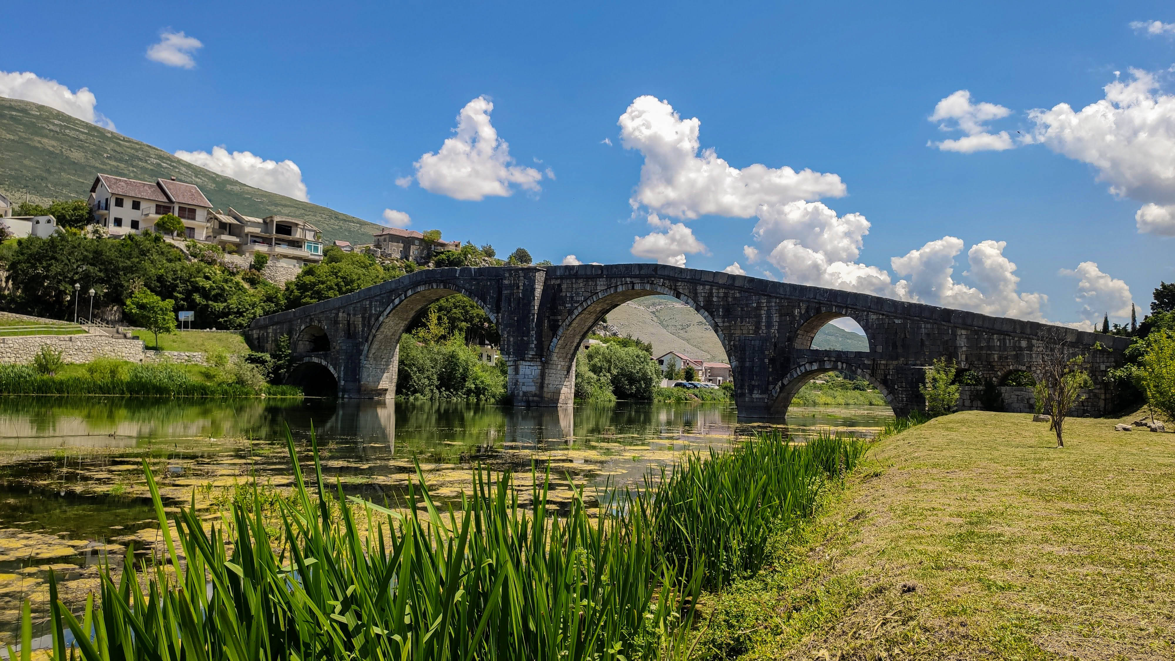 Bosnia And Herzegovina 桌布 壁紙 — 古老石橋有多個拱門，橫跨一條平靜的河流，周圍是綠色的蘆葦和山丘，在明亮的藍天白雲下。
