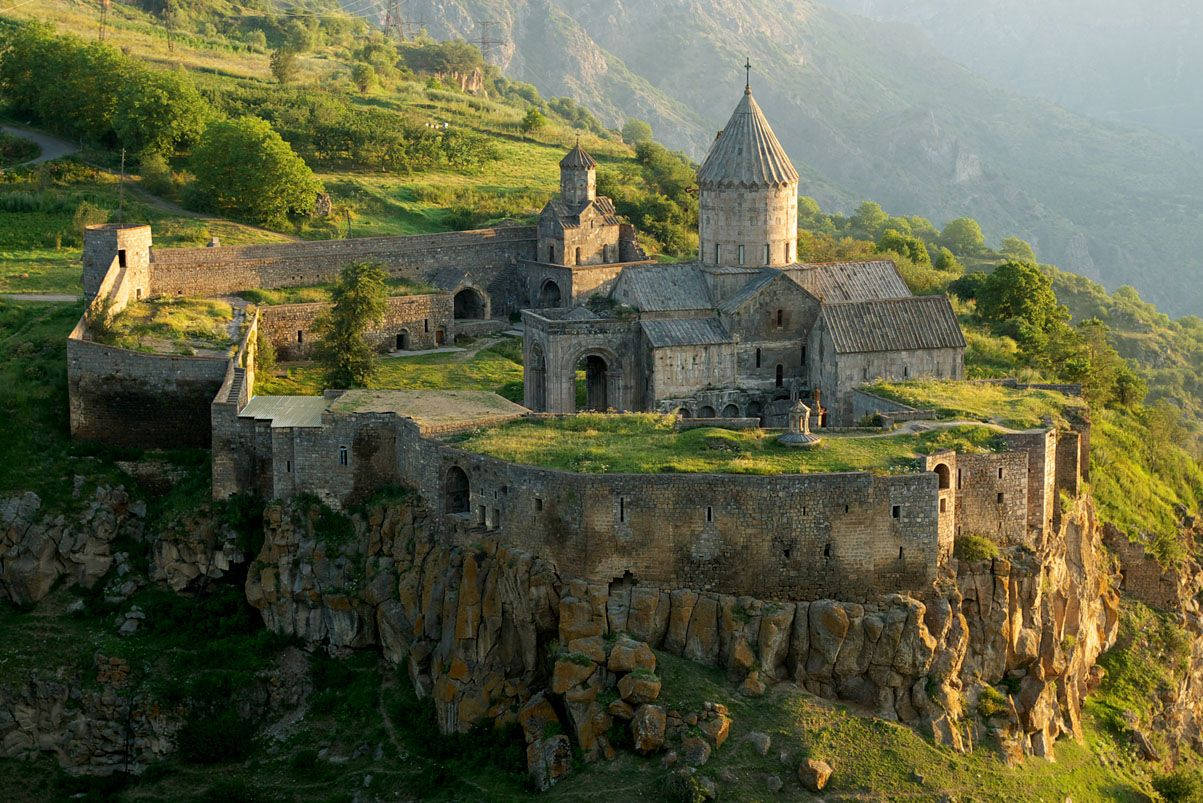 Armenia 桌布 壁紙 — 古老石造修道院坐落在崎嶇懸崖上，沐浴在金色日出光芒中，背景是鬱鬱蔥蔥的綠色山脈。
