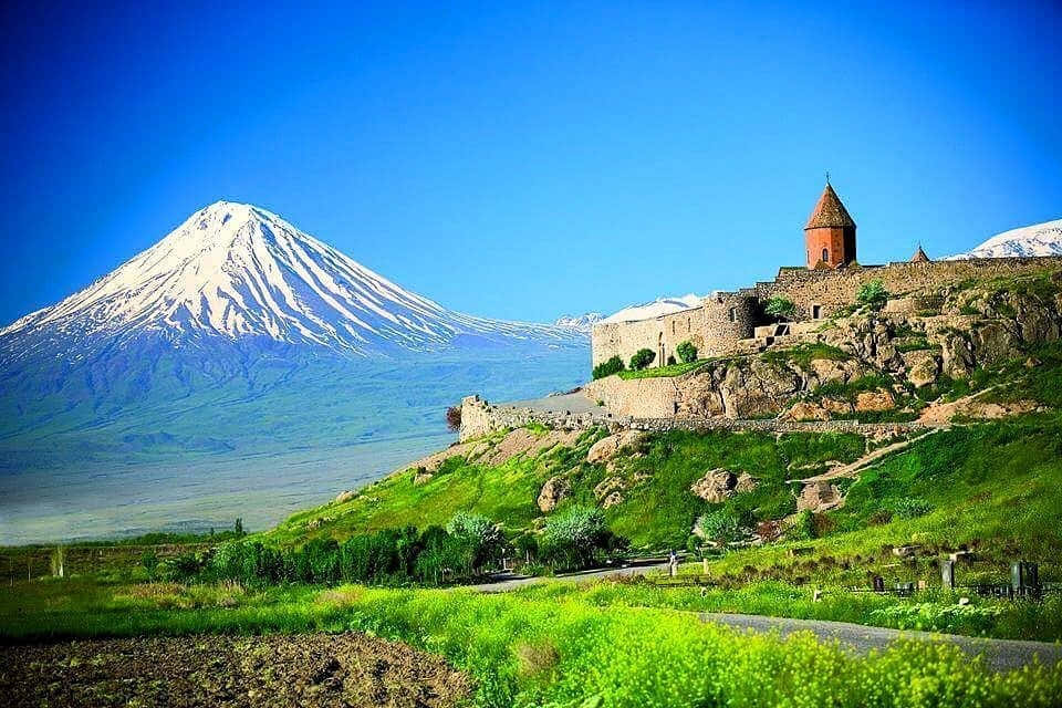 風景 桌布 壁紙 — 古老石造修道院坐落在綠色山坡上，背景中一座白雪覆蓋的山峰在清澈藍天下雄偉地聳立。