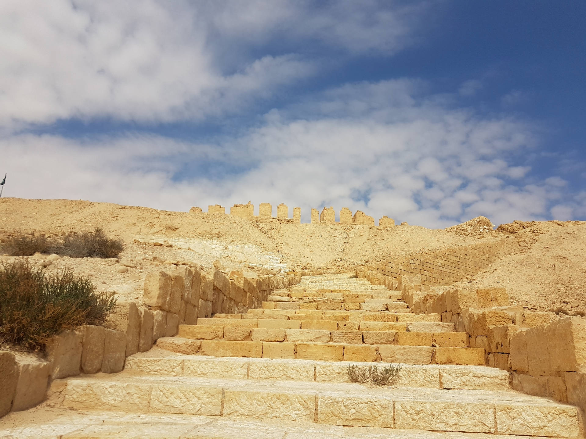 Israel 桌布 壁紙 — 古老石階在藍天白雲下，沿著烈日曝曬的沙漠山丘向上延伸，通往 crumbling 的堡壘城牆。
