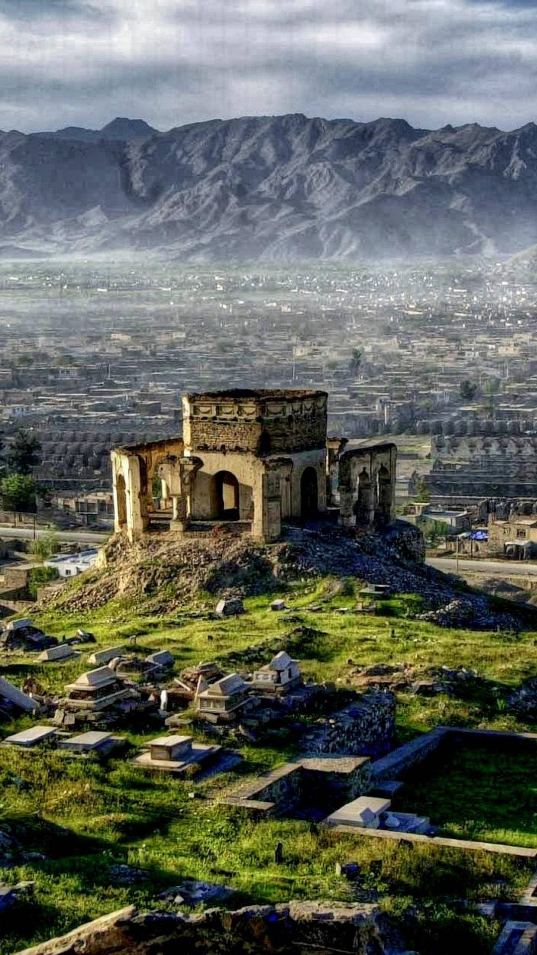Afghanistan 桌布 壁紙 — 古老石頭廢墟坐落在長滿草的山丘上，俯瞰著山谷中一座蔓延的城市，背景是霧濛濛的山脈。