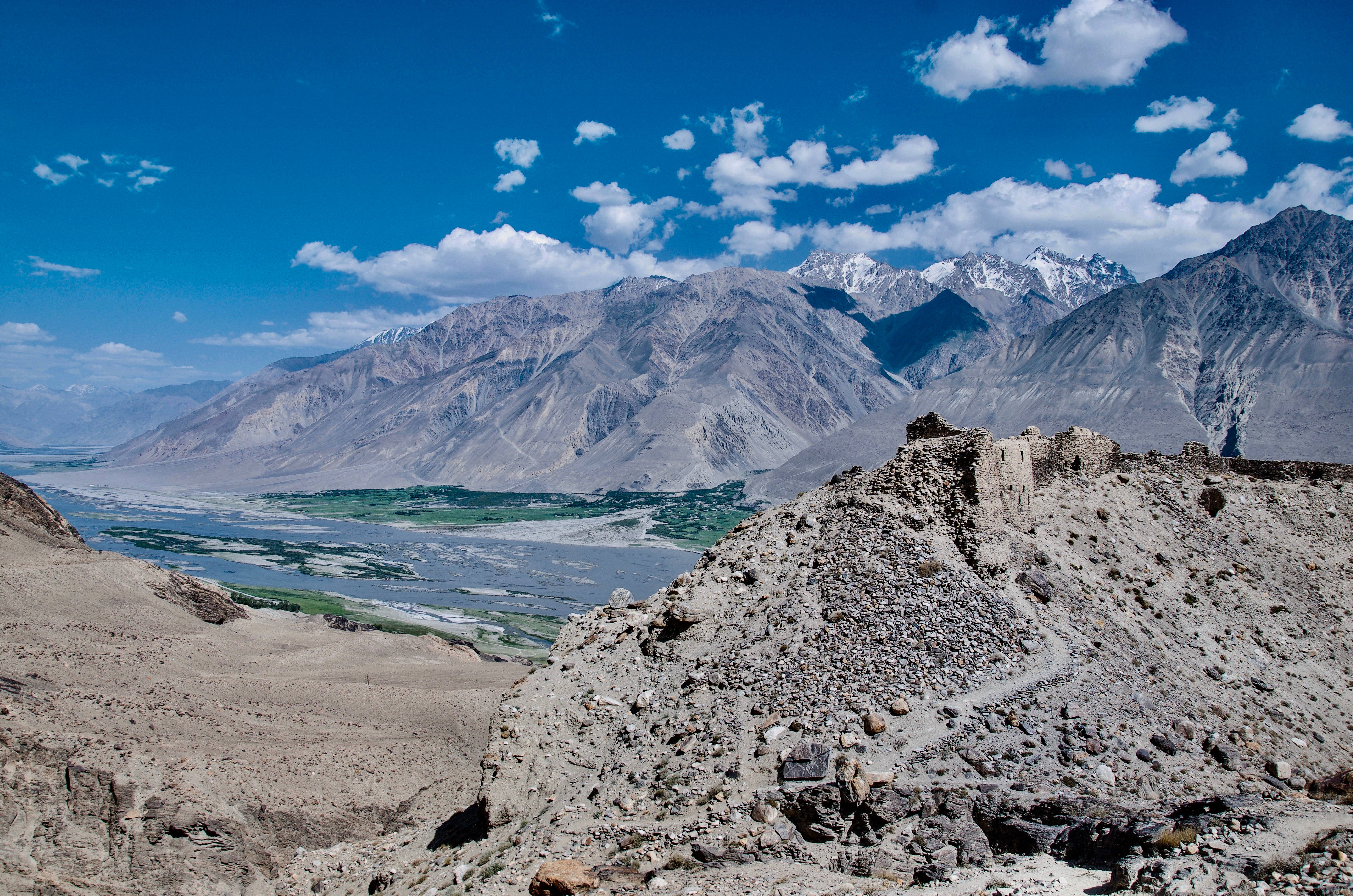 Tajikistan 桌布 壁紙 — 古老石頭廢墟棲息在岩石山脊頂端，俯瞰蜿蜒的河谷，兩側是白雪覆蓋的山峰，上方是明亮的藍天，點綴著散落的雲朵。