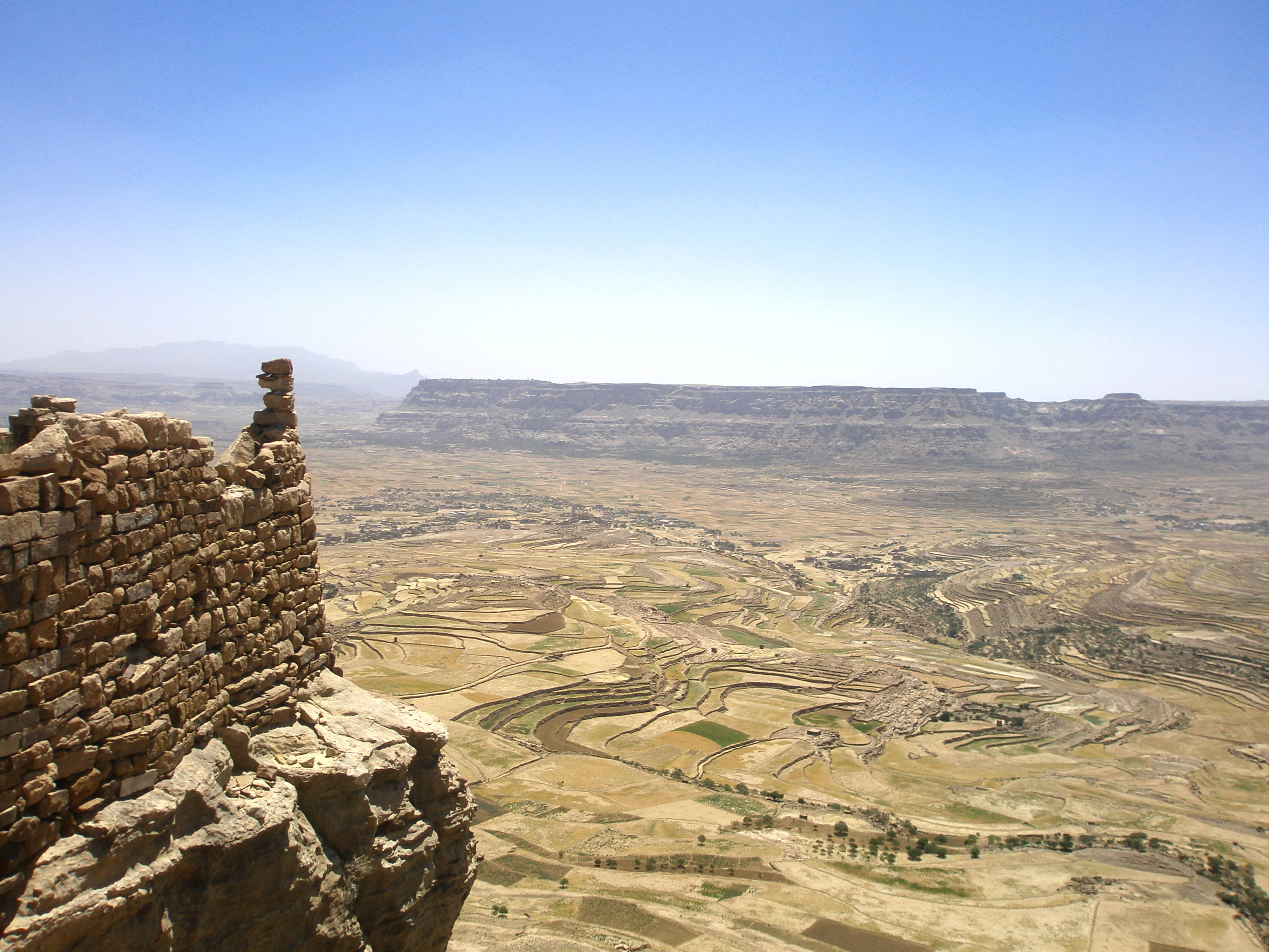 Yemen 桌布 壁紙 — 古老石頭遺跡俯瞰廣闊的梯田山谷，天空湛藍清澈，遠山連綿。