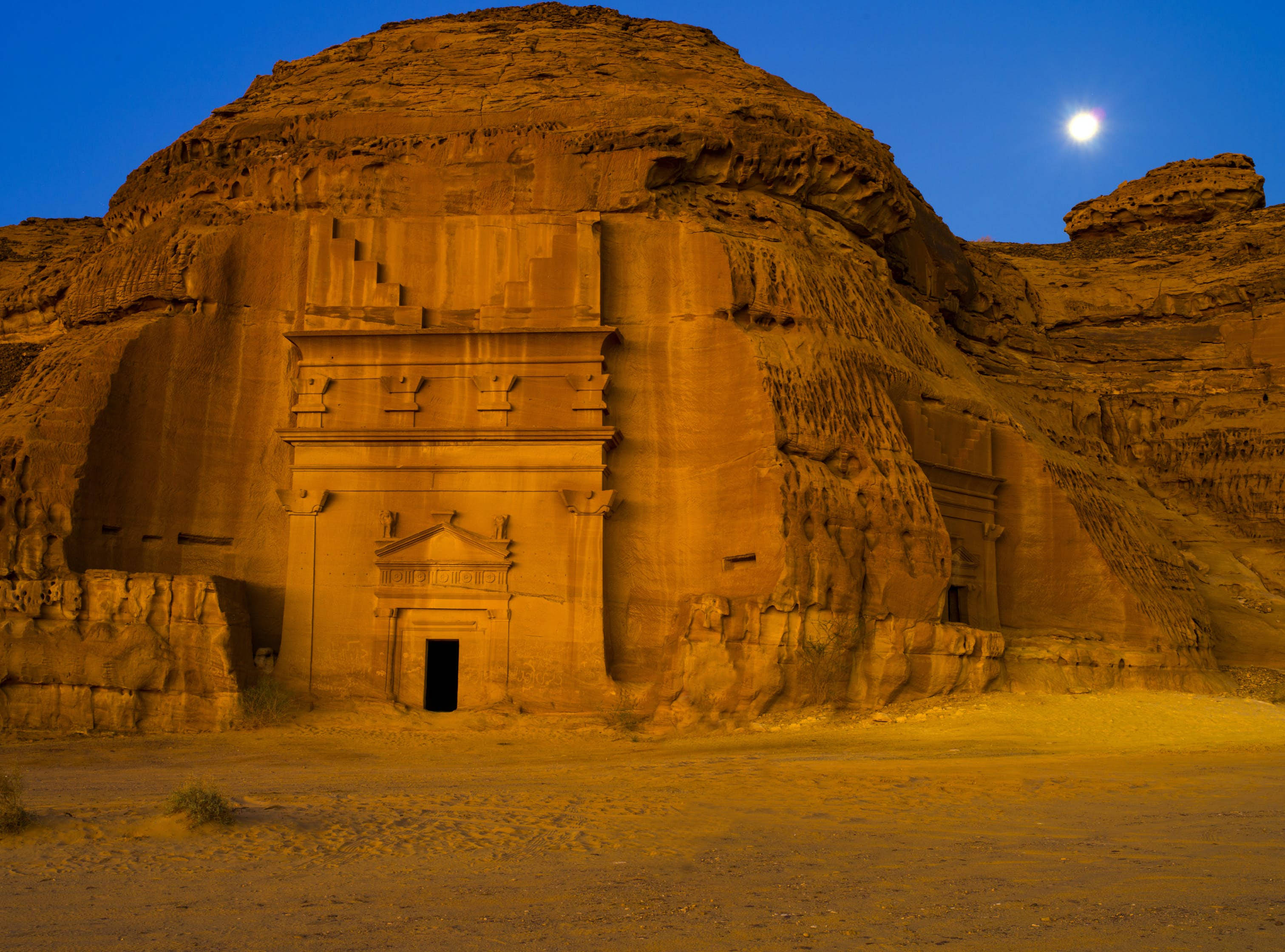 Saudi 桌布 壁紙 — 古老雕刻寺廟外牆在月光下閃耀琥珀色光芒，映襯著深藍色的沙漠夜空。