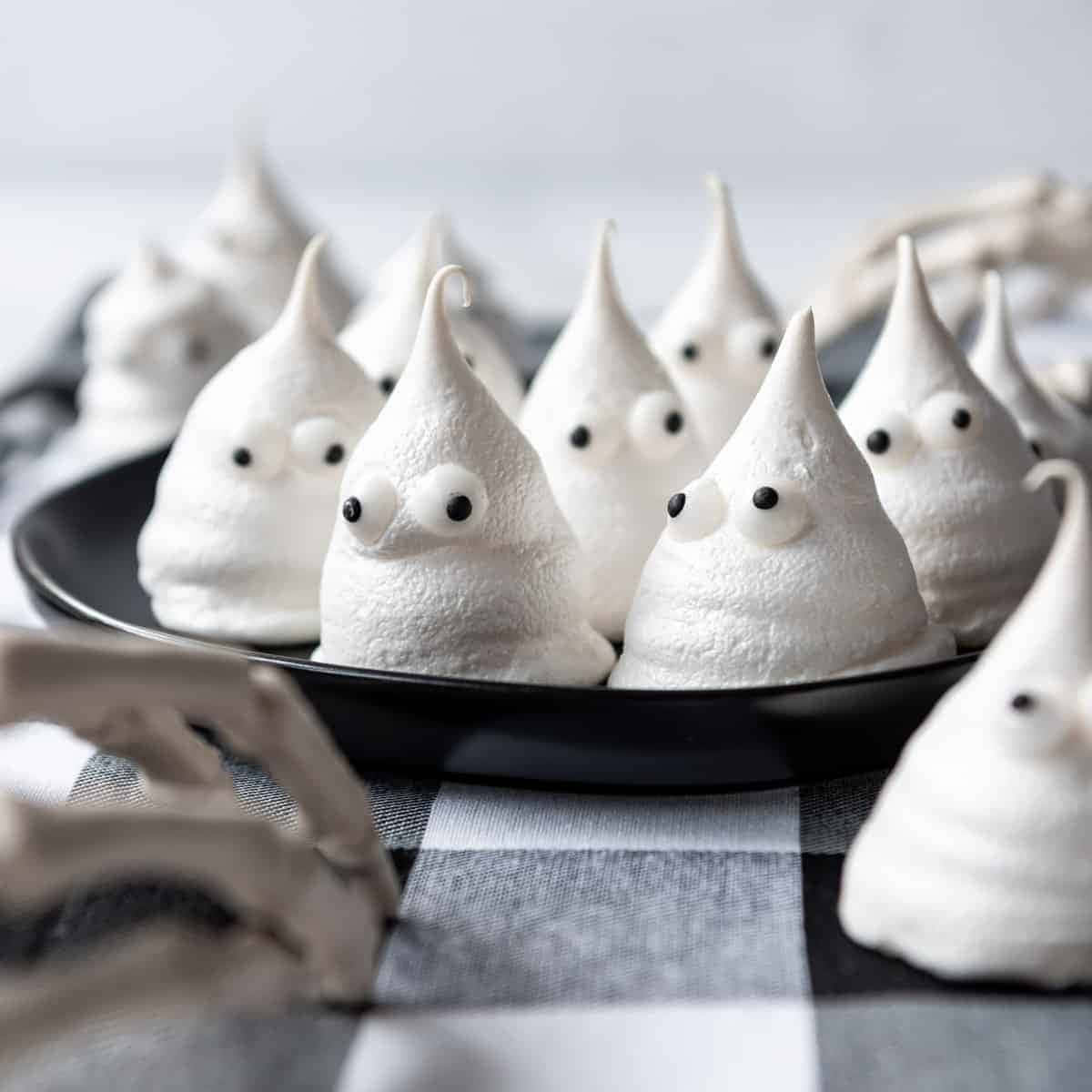 Meringue 桌布 壁紙 — 可愛的白色蛋白霜鬼魂，帶有咕嚕眼睛，坐在黑色盤子上，盤子放在格紋布上，附近有骷髏手。