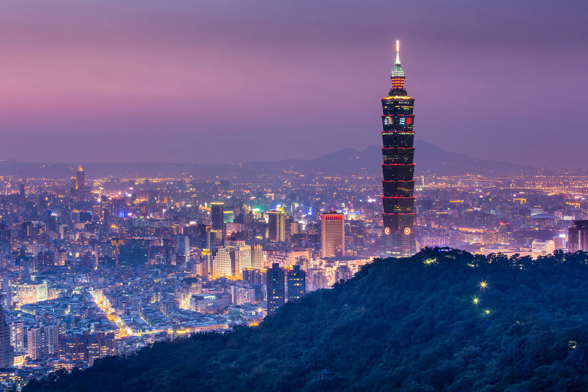 台北101在黃昏時亮起，俯瞰密集的城市，天空呈紫色，山丘翠綠。