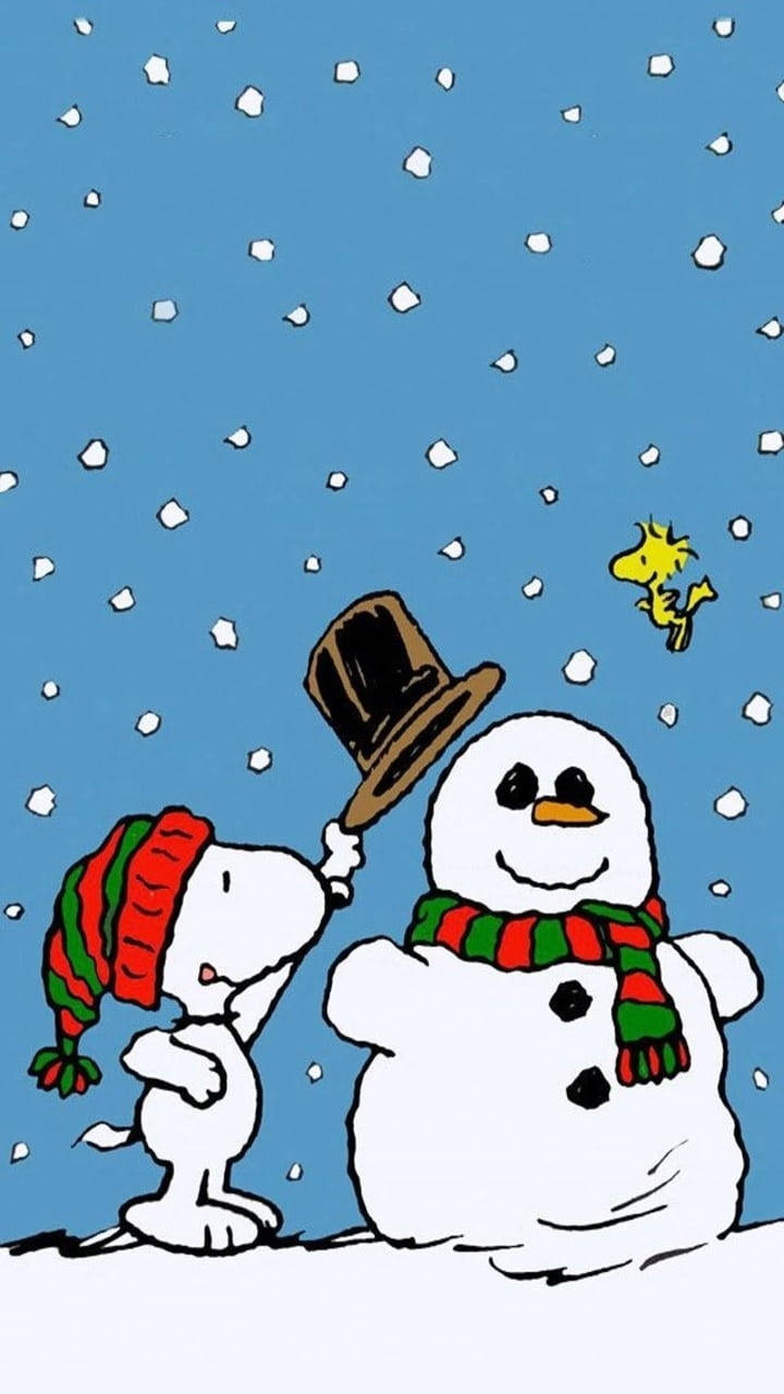 Snoopy Christmas 桌布 壁紙 — 史努比戴著紅綠色的帽子，輕輕地將一頂高帽放在雪人上，而胡士托則在附近飛翔，背景是雪白的藍天。