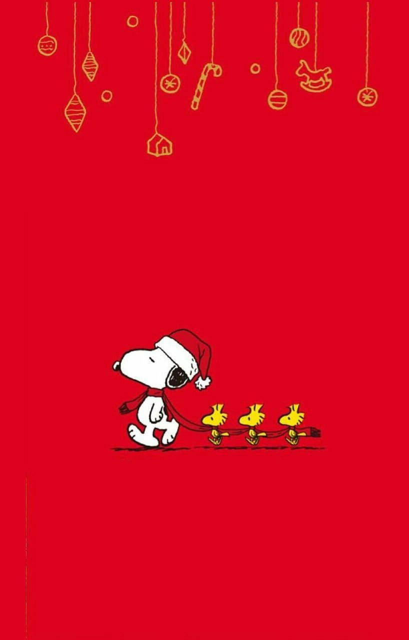 Snoopy Christmas 桌布 壁紙 — 史努比戴著聖誕帽，在掛有金色裝飾的鮮紅色背景前，用皮帶拉著三隻黃色小雞。