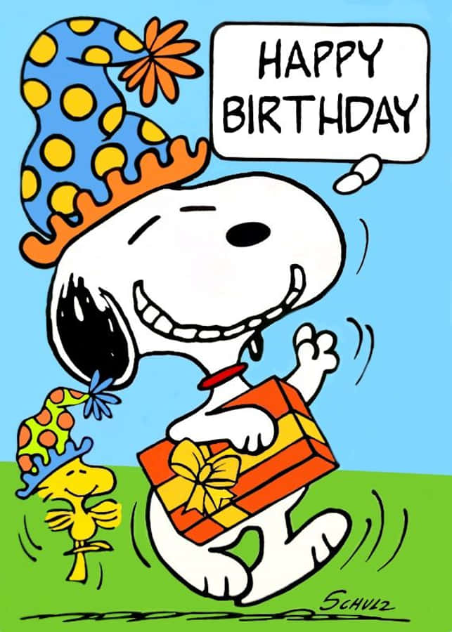 Charlie Brown Birthday 桌布 壁紙 — 史努比戴著藍色圓點派對帽，高興地拿著一個繫有黃色蝴蝶結的禮物盒，而胡士托在他旁邊，在「生日快樂」對話框下跳舞。