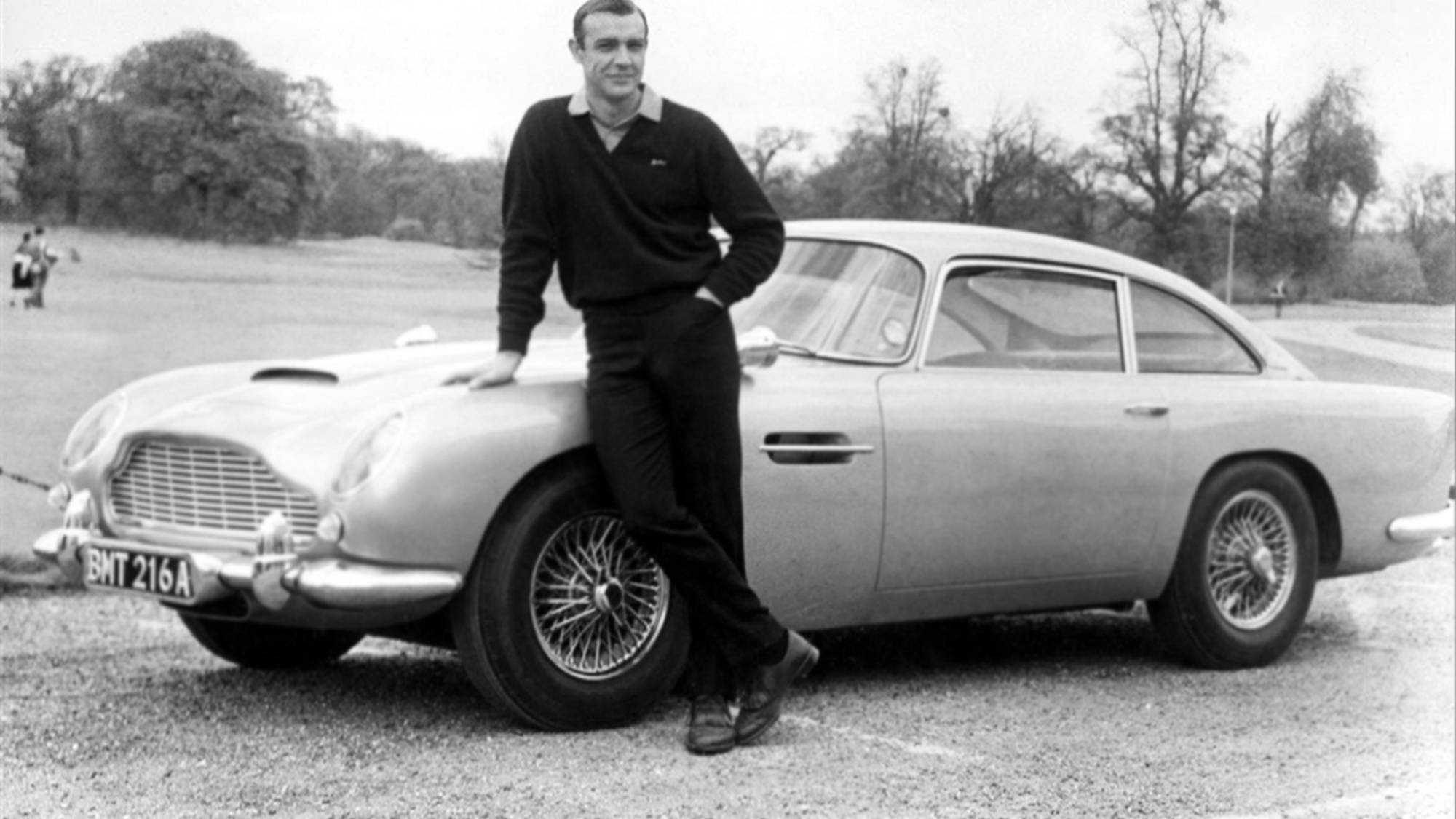 Sean Connery 桌布 壁紙 — 史恩·康納萊在碎石路上休閒地靠著經典銀色Aston Martin DB5，背景有樹木。