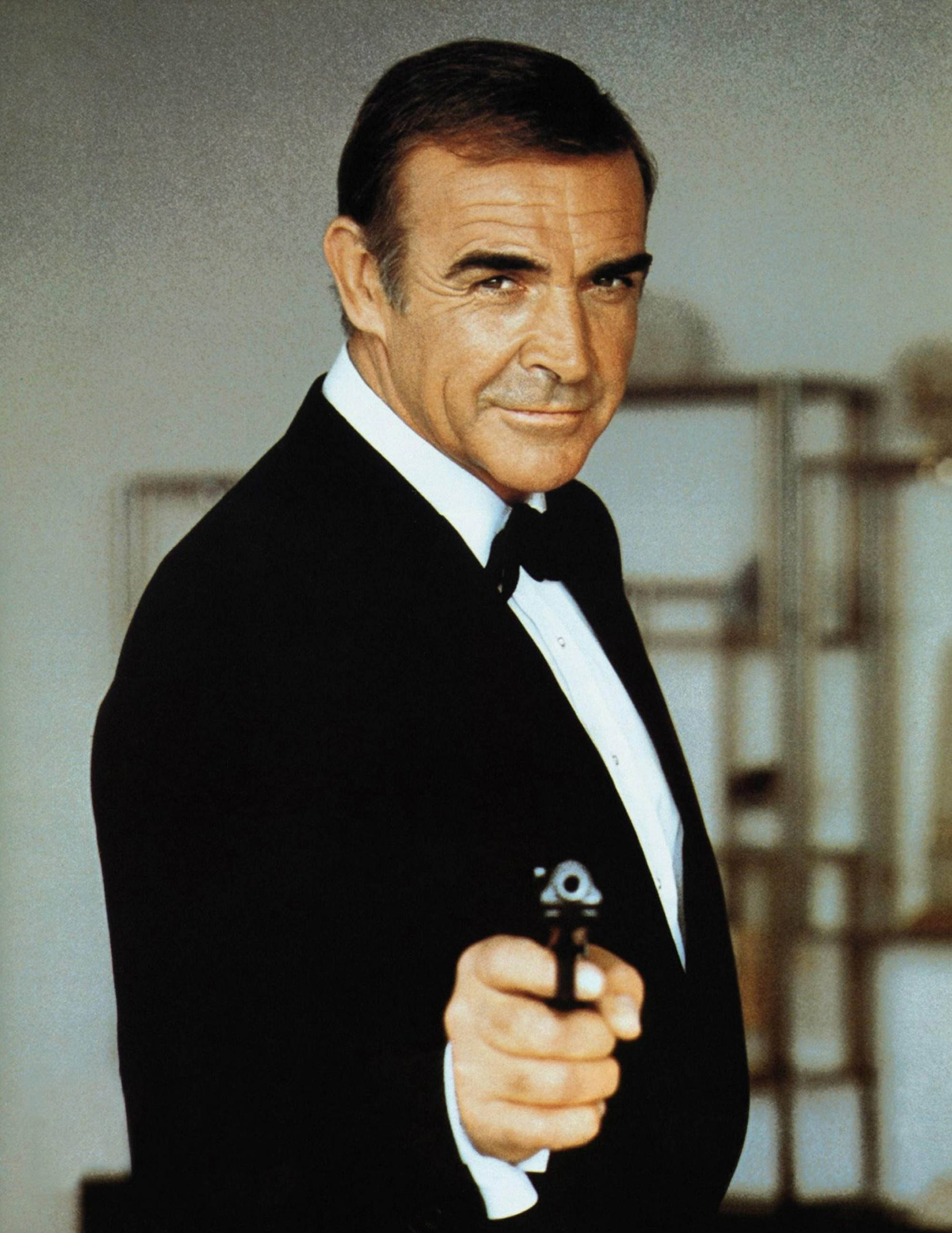 Sean Connery 桌布 壁紙 — 史恩·康納萊飾演詹姆士·龐德，身穿禮服，手持手槍，自信微笑，背景為模糊室內。