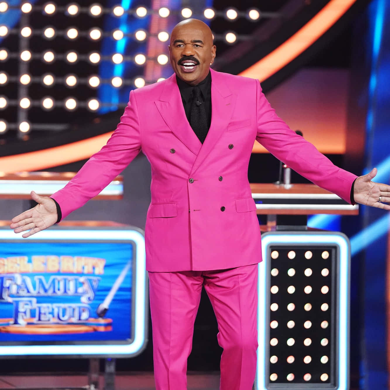 steve harvey 桌布 壁紙 — 史提夫·哈維穿著鮮豔的粉紅色西裝，站在遊戲節目舞台上，張開雙臂，表情喜悅。
