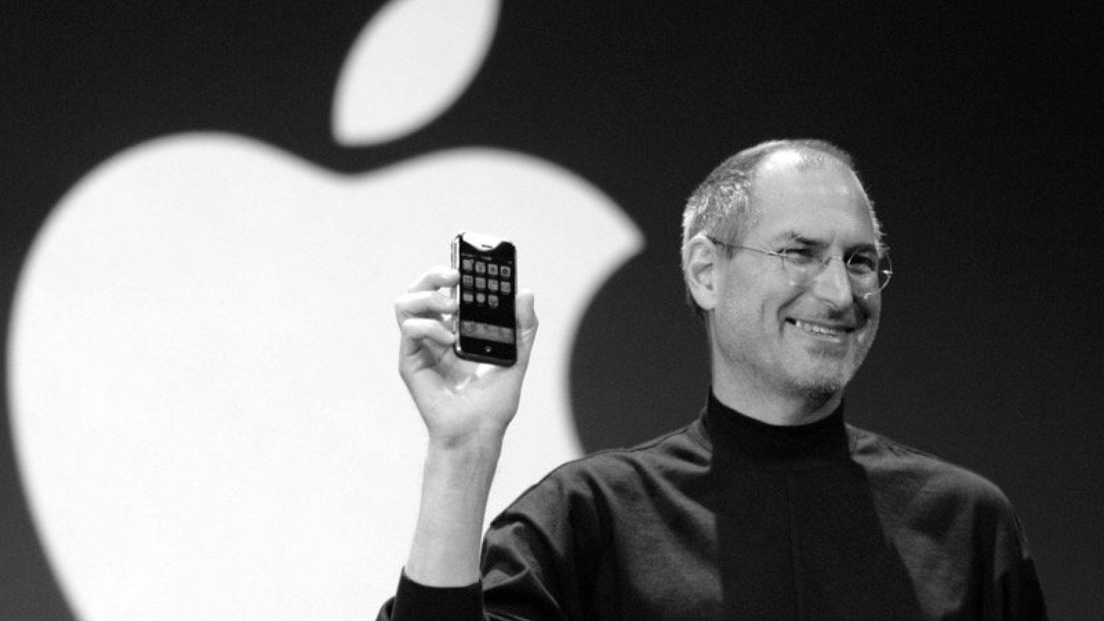 Rich 桌布 壁紙 — 史提夫·賈伯斯微笑著舉起一部早期iPhone，背景是巨大的黑白Apple標誌。