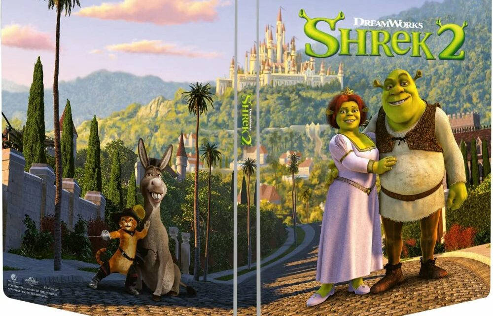 Shrek 2 桌布 壁紙 — 史瑞克、費歐娜、驢子、鞋貓在粉紅天空下的城堡前，站在鵝卵石小徑上微笑，背景有棕櫚樹和山脈。