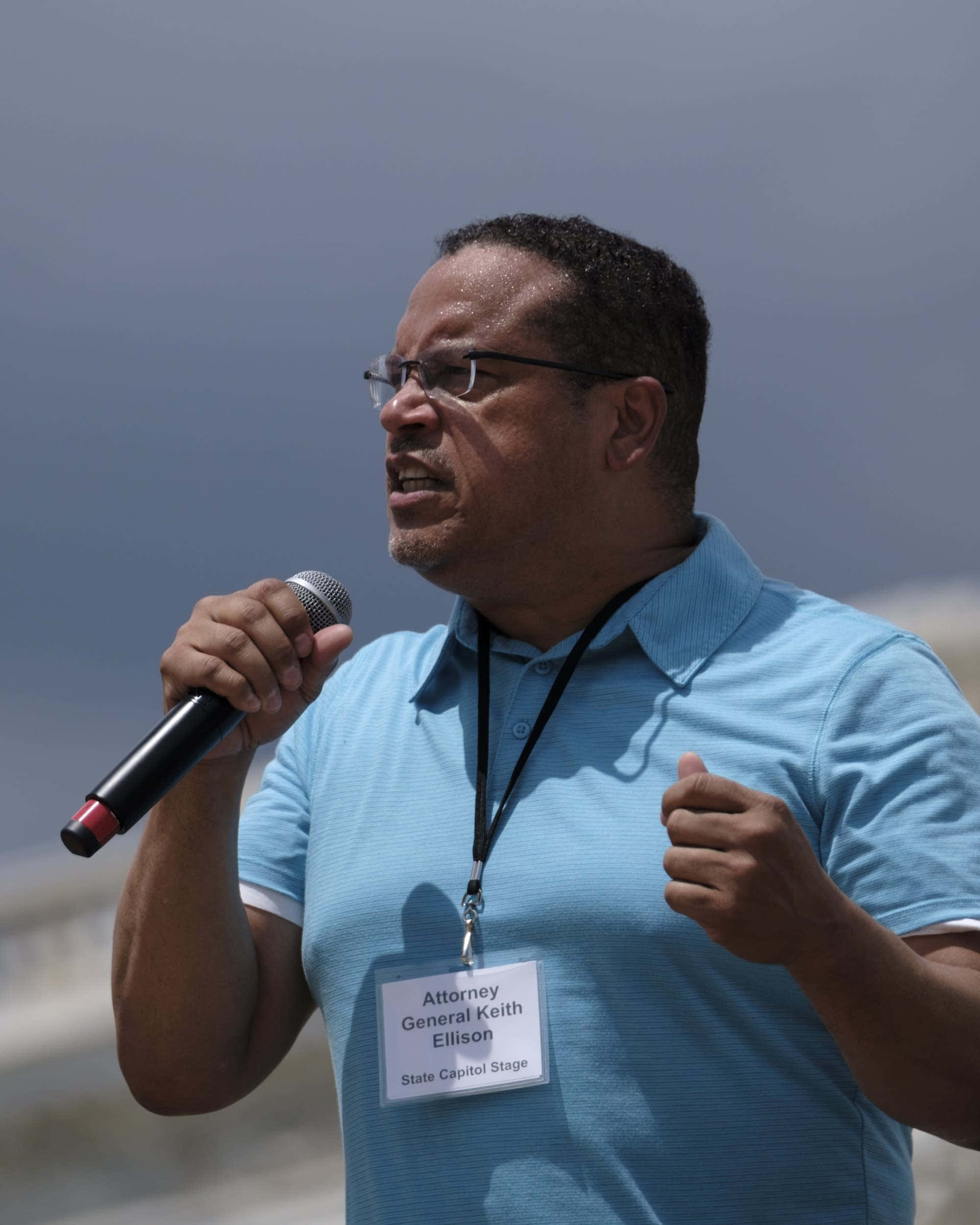 Microphone 桌布 壁紙 — 司法部長 Keith Ellison 在日間戶外活動中，穿著淺藍色 Polo 衫，熱情地對著麥克風講話。