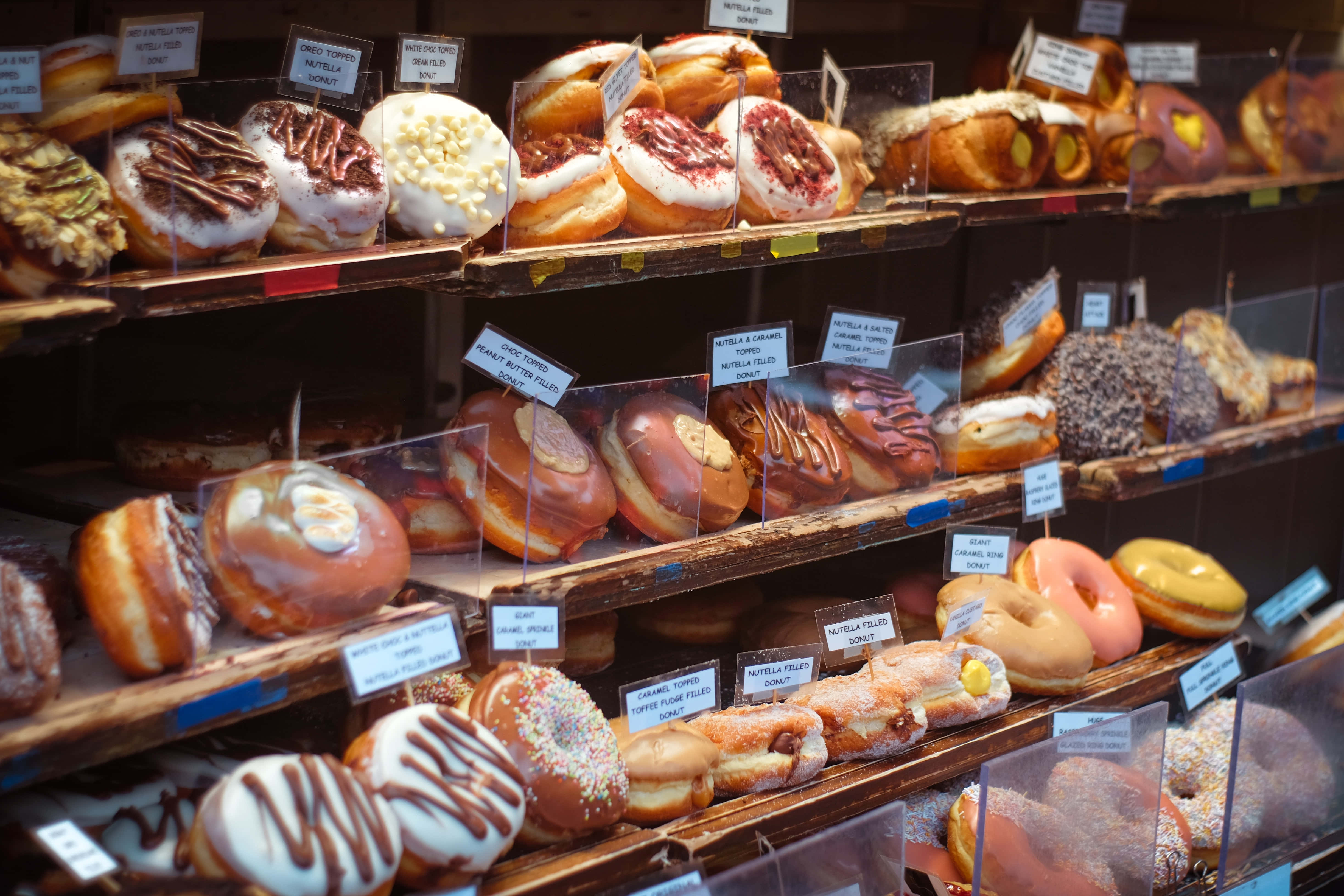 Bakery 桌布 壁紙 — 各式各樣有糖霜和配料的甜甜圈整齊排列在麵包店陳列櫃中，附有價格標籤。